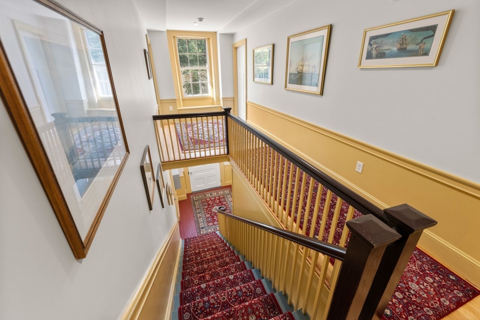 Carpeted staircase with dark wood railings, yellow wainscoting, framed artwork, and a bright window.