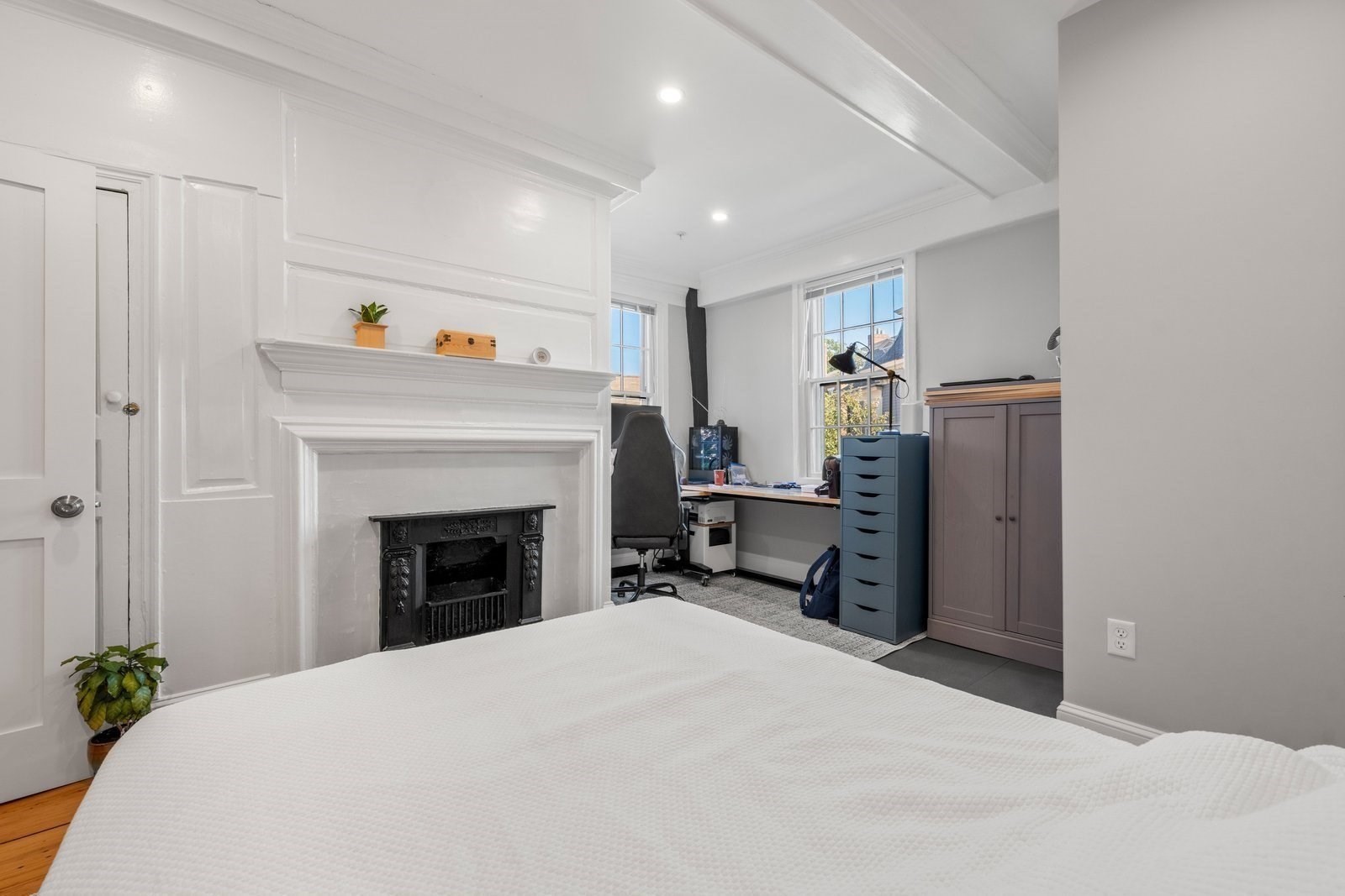 Bedroom with an ornate white fireplace, black insert, and desk beside two windows.