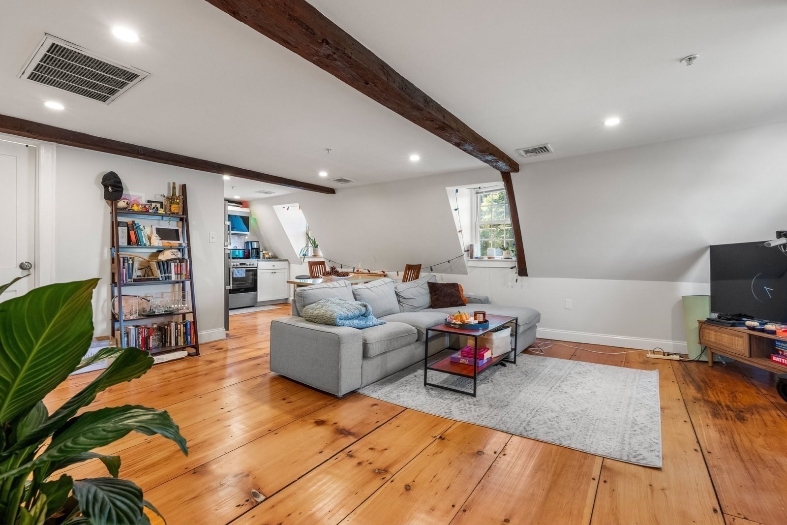 Living room with exposed beams, wide-plank wood floors, gray sectional, and open kitchen.