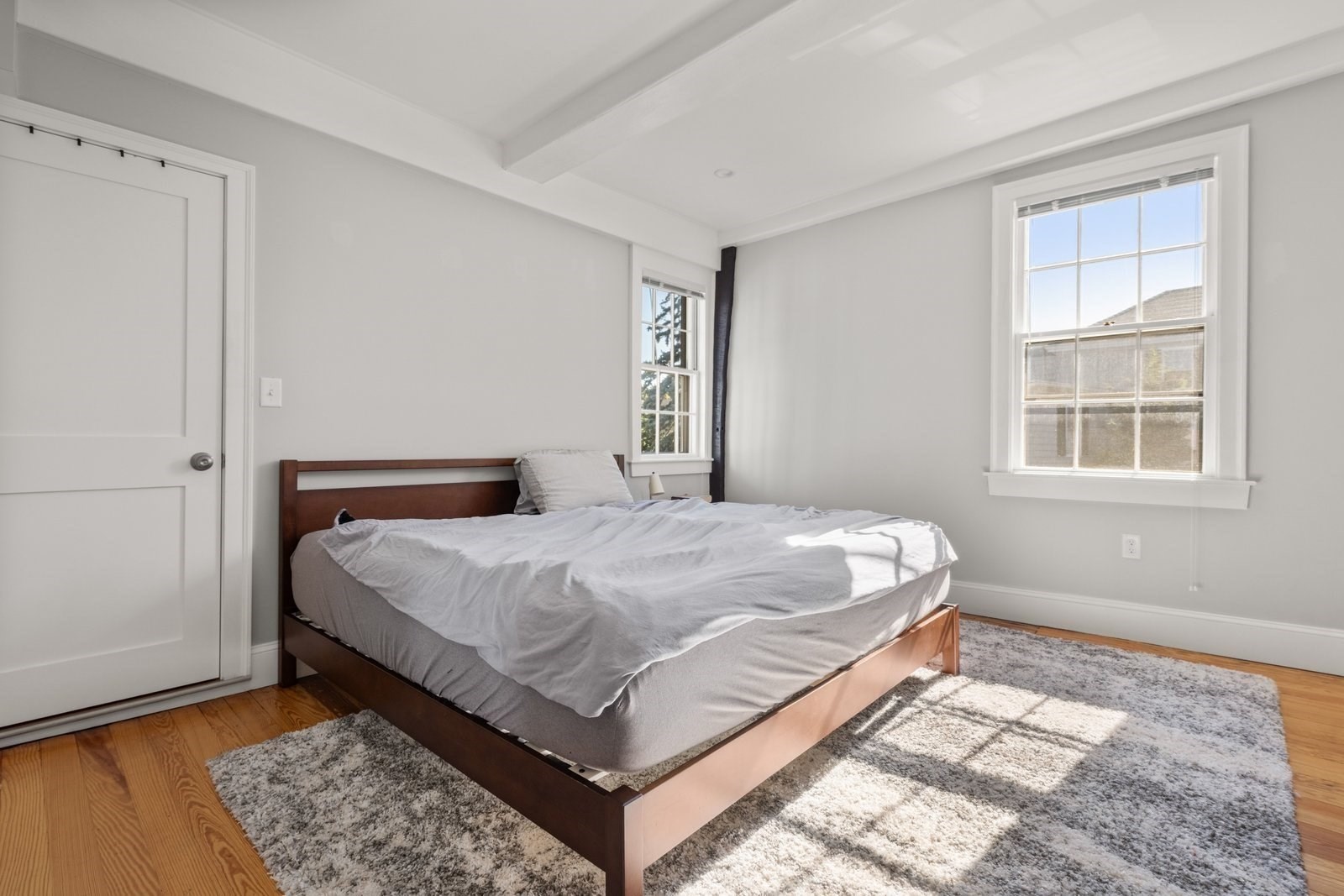 Bedroom with a wooden bed, gray bedding, two sunlit windows, and hardwood floors.