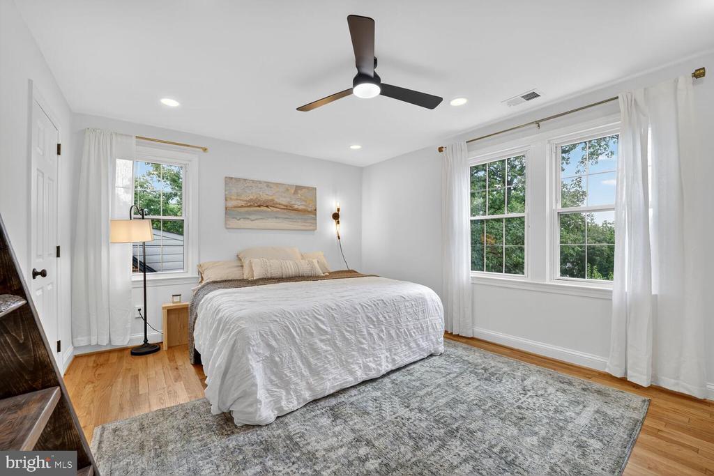 Furnished bedroom with a white bed, gray rug, ceiling fan, and large windows.