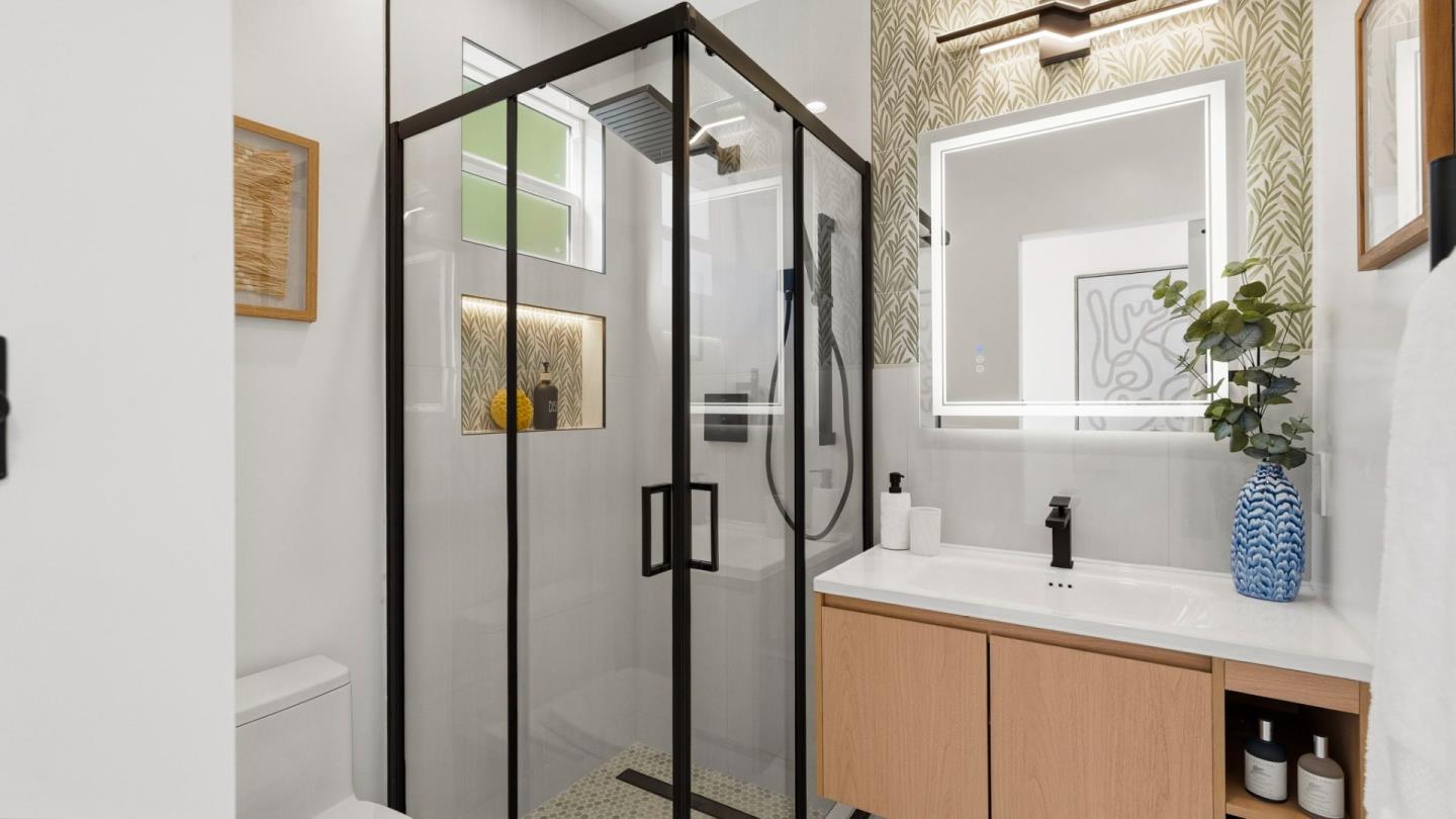 Bathroom with a black-framed glass shower, light wood vanity, and leafy patterned wallpaper.
