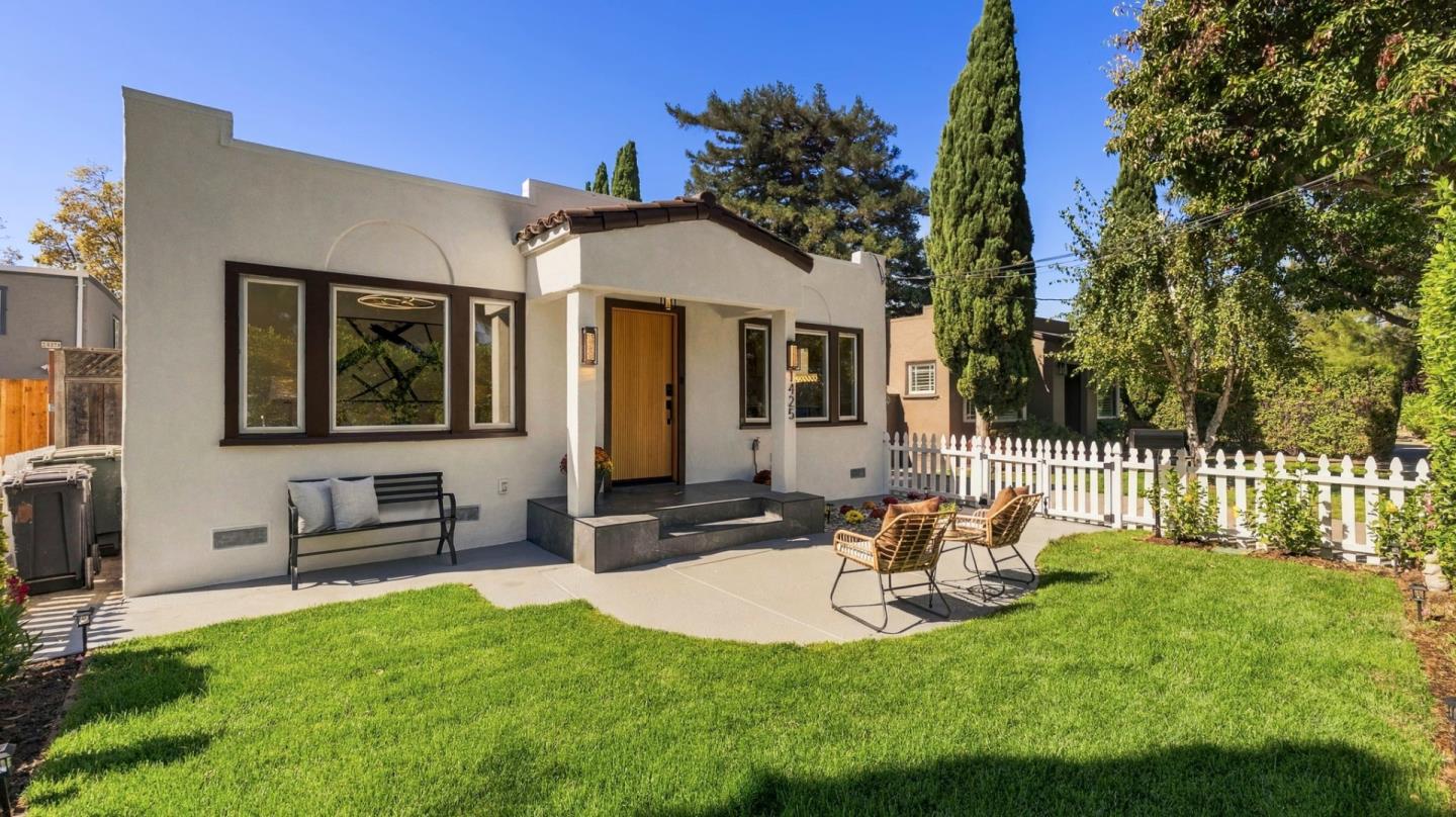 White stucco home with a wood front door, lawn, patio chairs, and white picket fence.