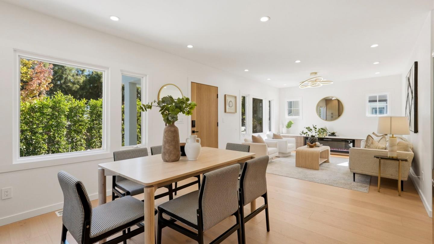Open-plan dining and living room with a large wood table, neutral seating, and windows overlooking greenery.