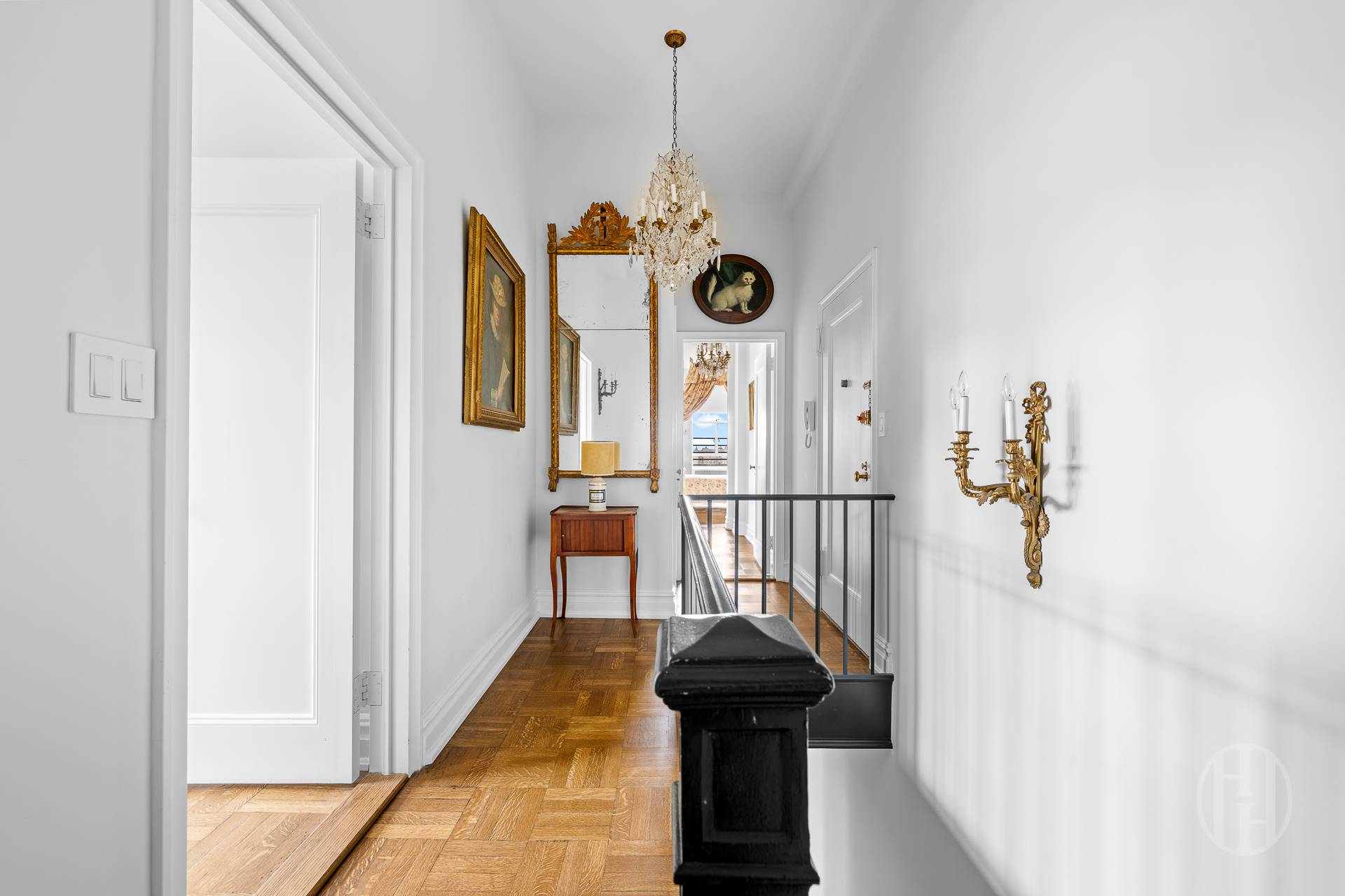 Bright apartment hallway with parquet floors, an ornate chandelier, gilt mirror, and black stair railing.