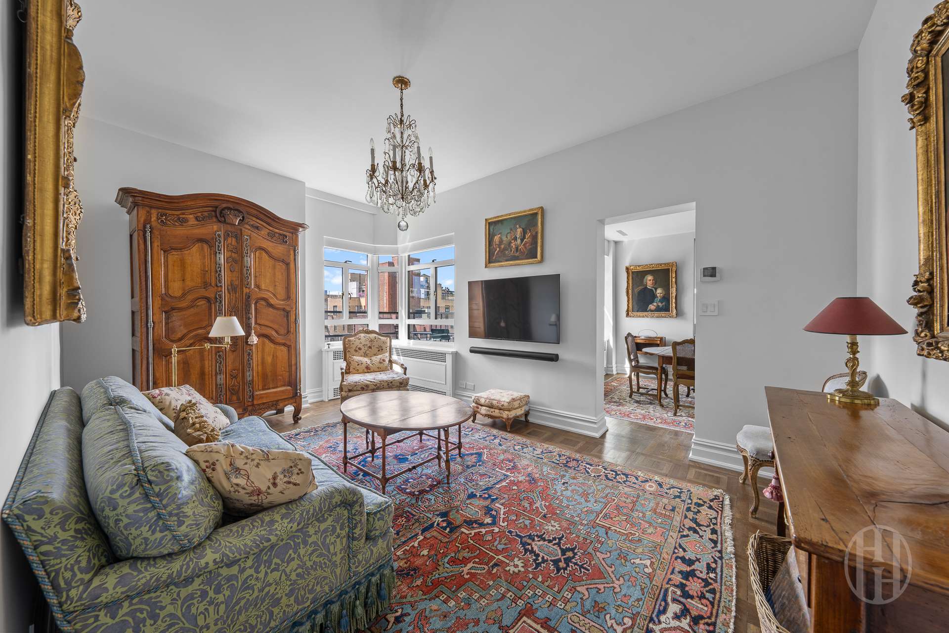 Living room with an ornate wooden armoire, crystal chandelier, and corner windows overlooking nearby buildings.