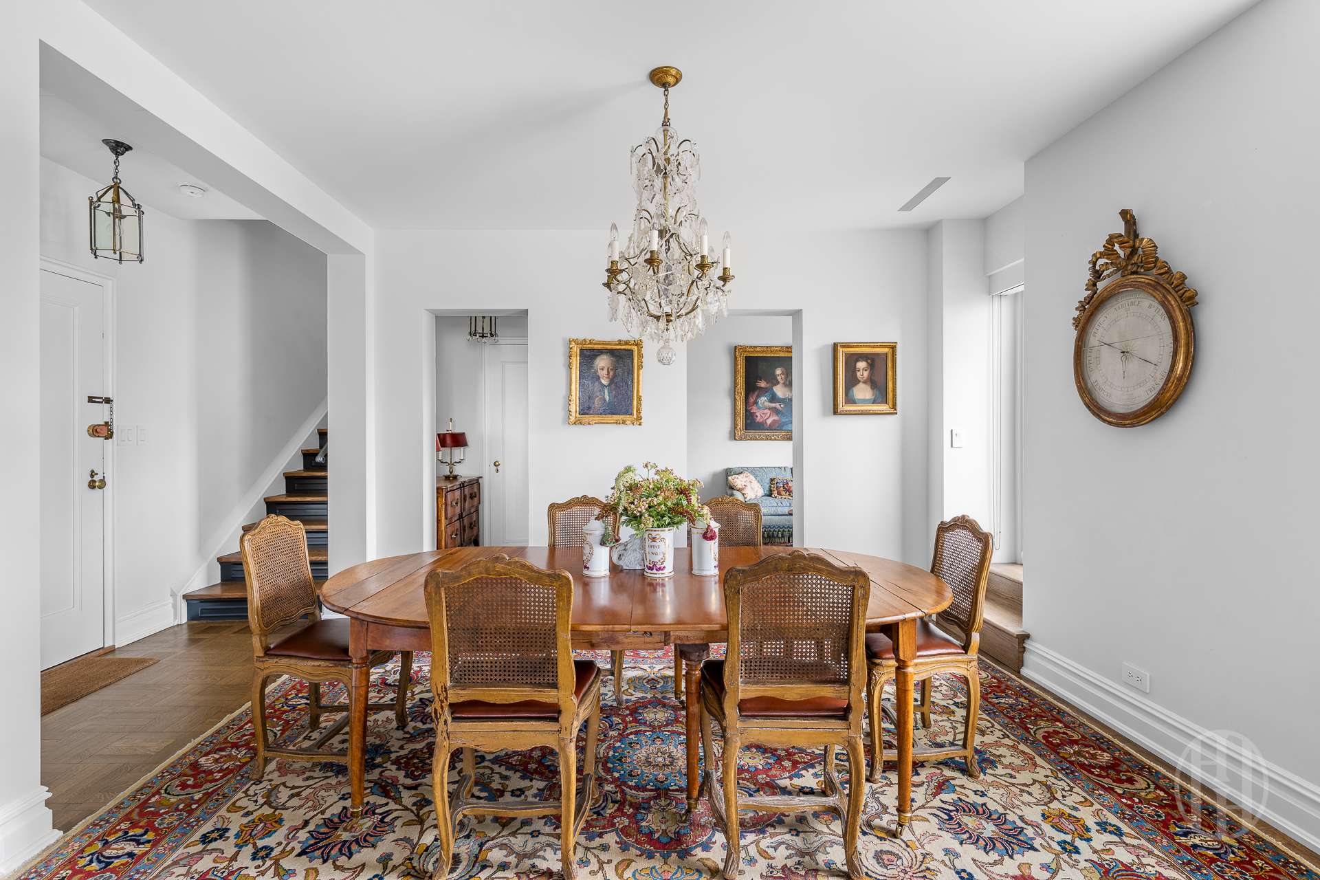 Dining room with an oval wooden table, crystal chandelier, patterned rug, and ornate wall clock.