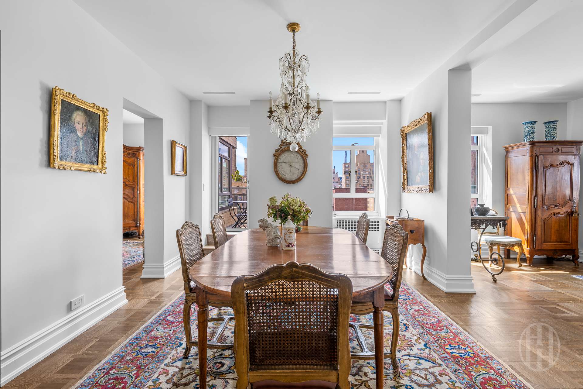 Furnished dining room with an antique wooden table, crystal chandelier, patterned rug, and city views through tall windows.