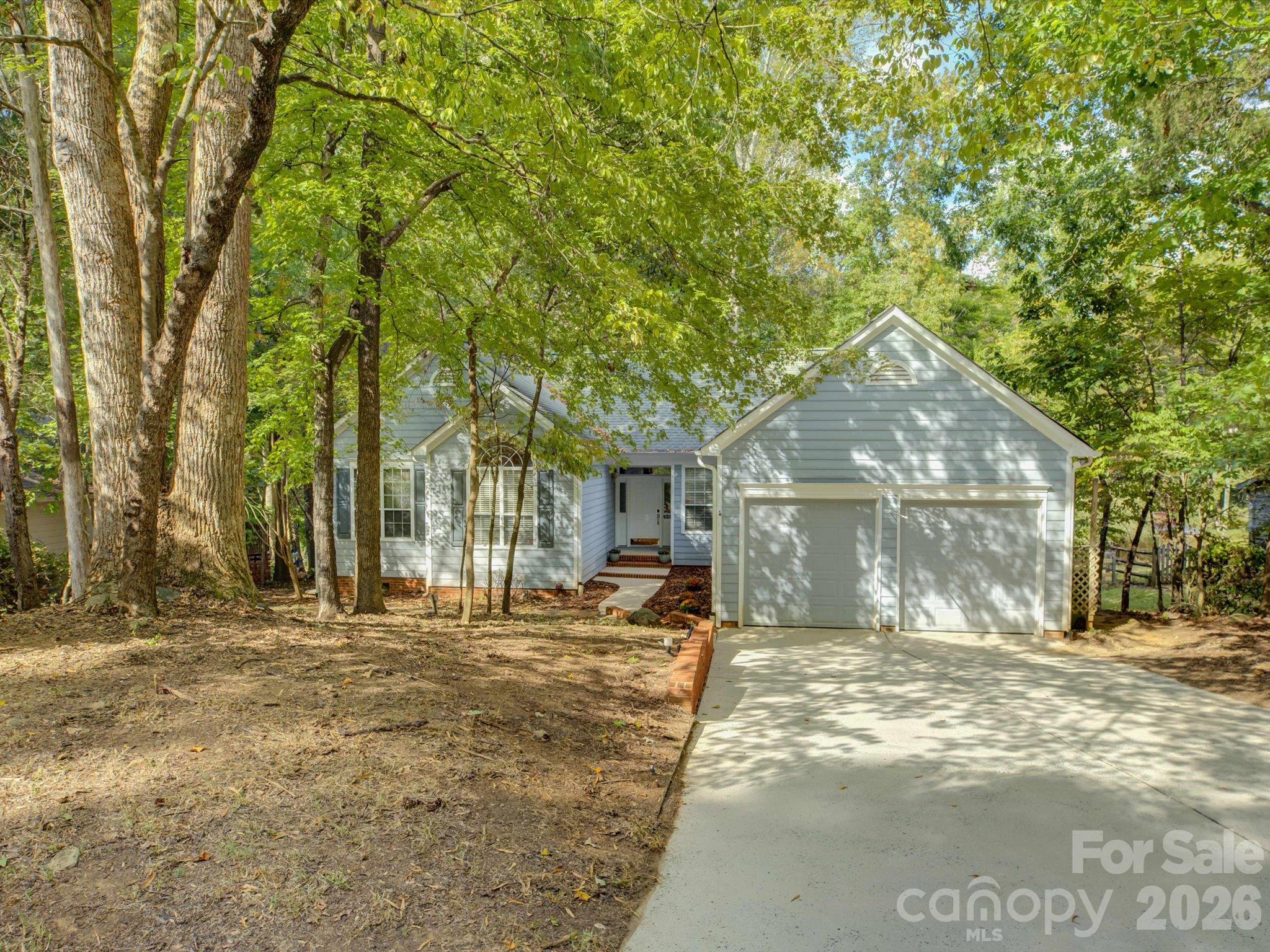 Light gray house with a two-car garage and driveway, surrounded by mature trees.