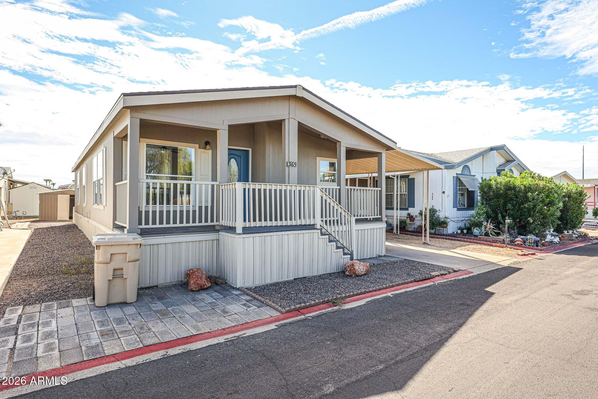 Raised manufactured home with a covered porch, white railings, and blue front door beside a paved lane.