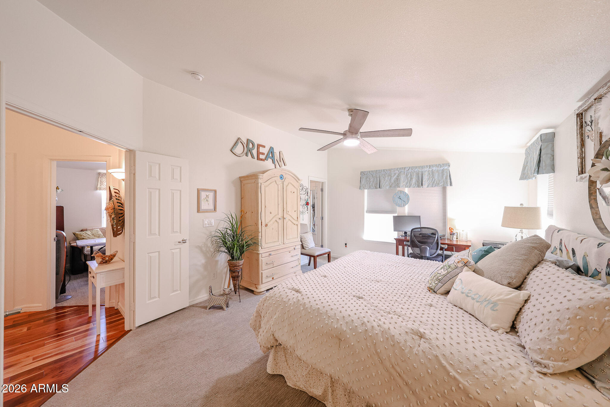 Furnished bedroom with a cream bed, ornate armoire, ceiling fan, and desk beside bright windows.