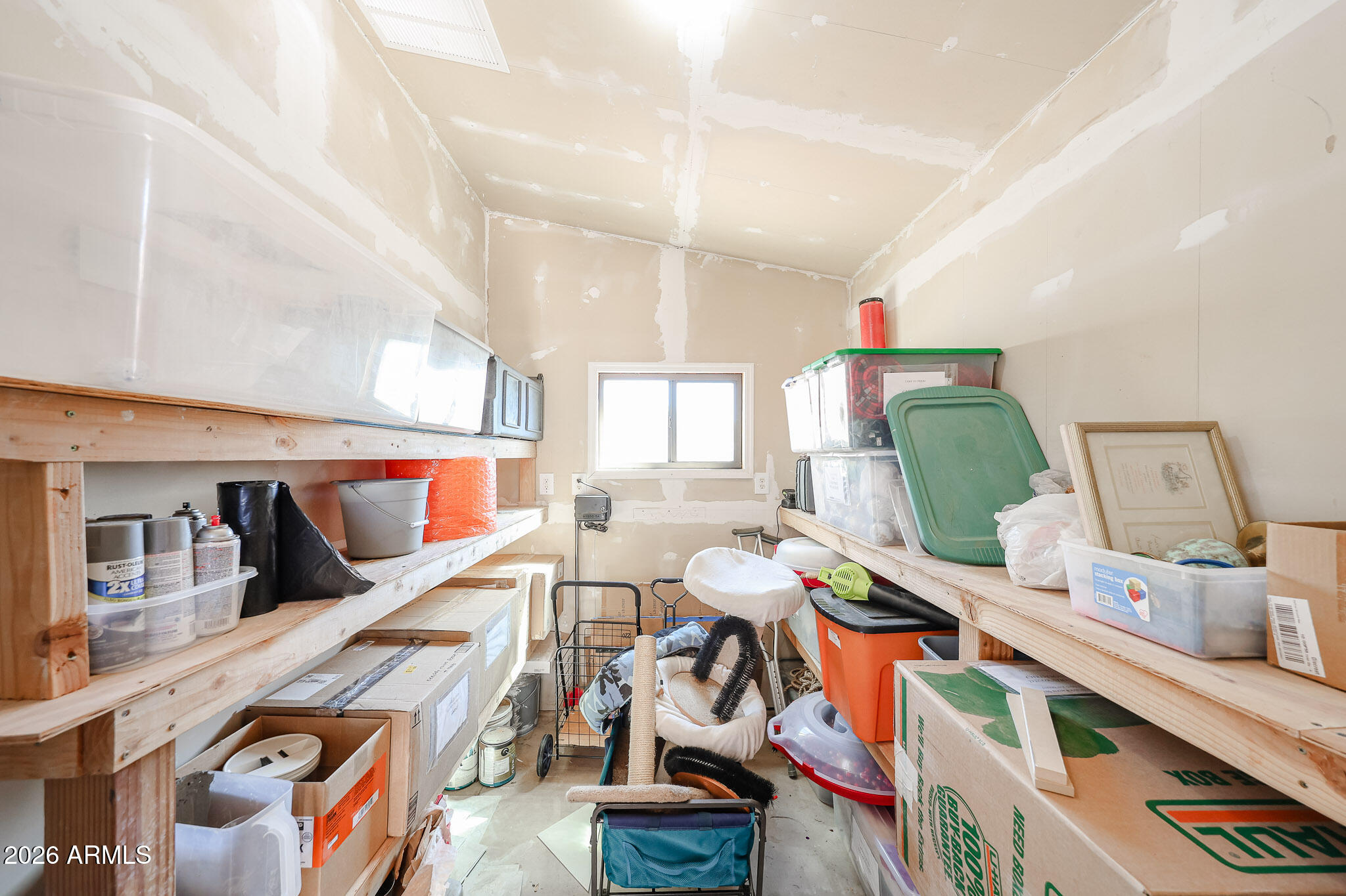 Crowded storage room with wooden shelves, stacked bins and boxes, and unfinished drywall.