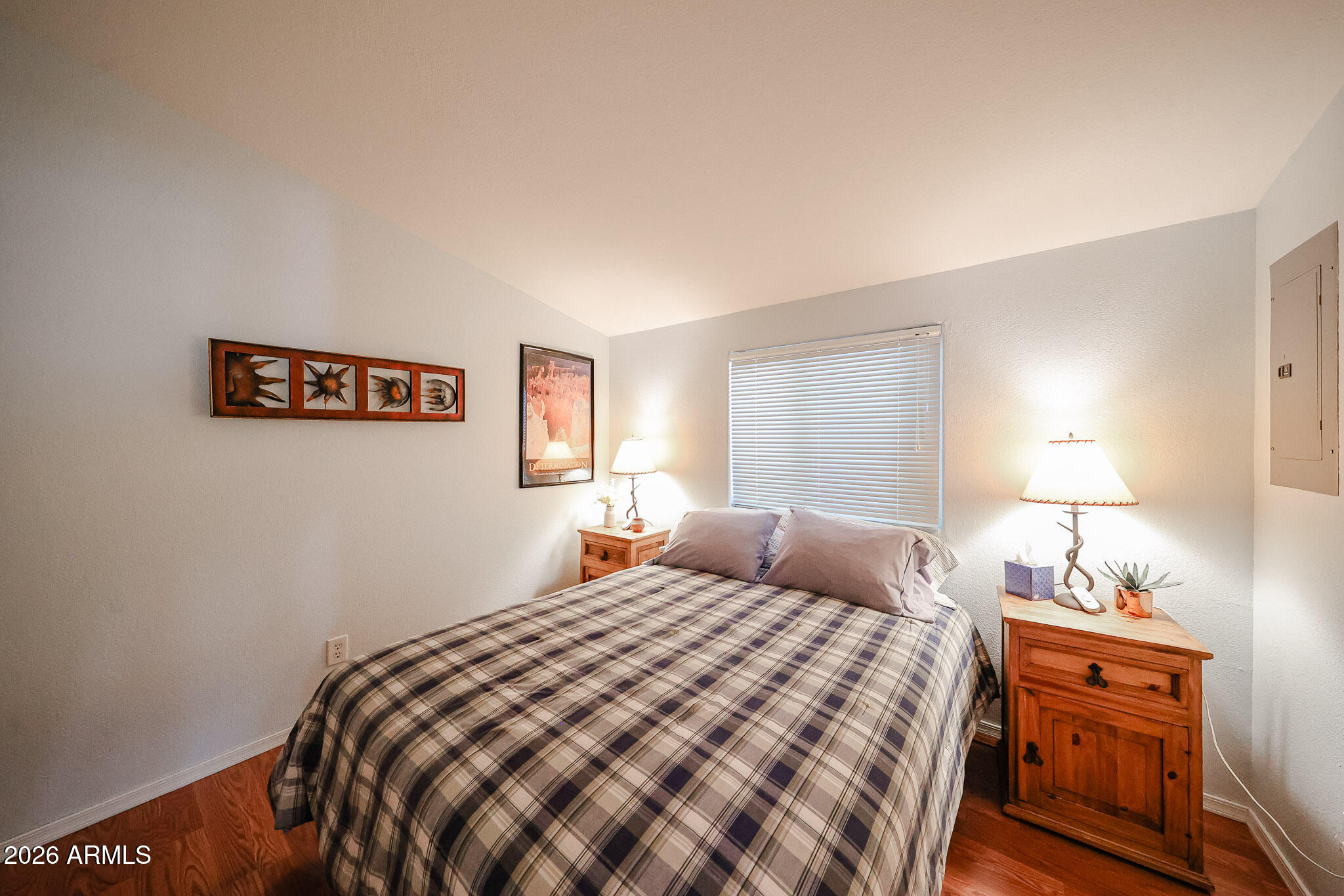 Bedroom with a plaid bedspread, wooden nightstands, lit lamps, and closed window blinds.