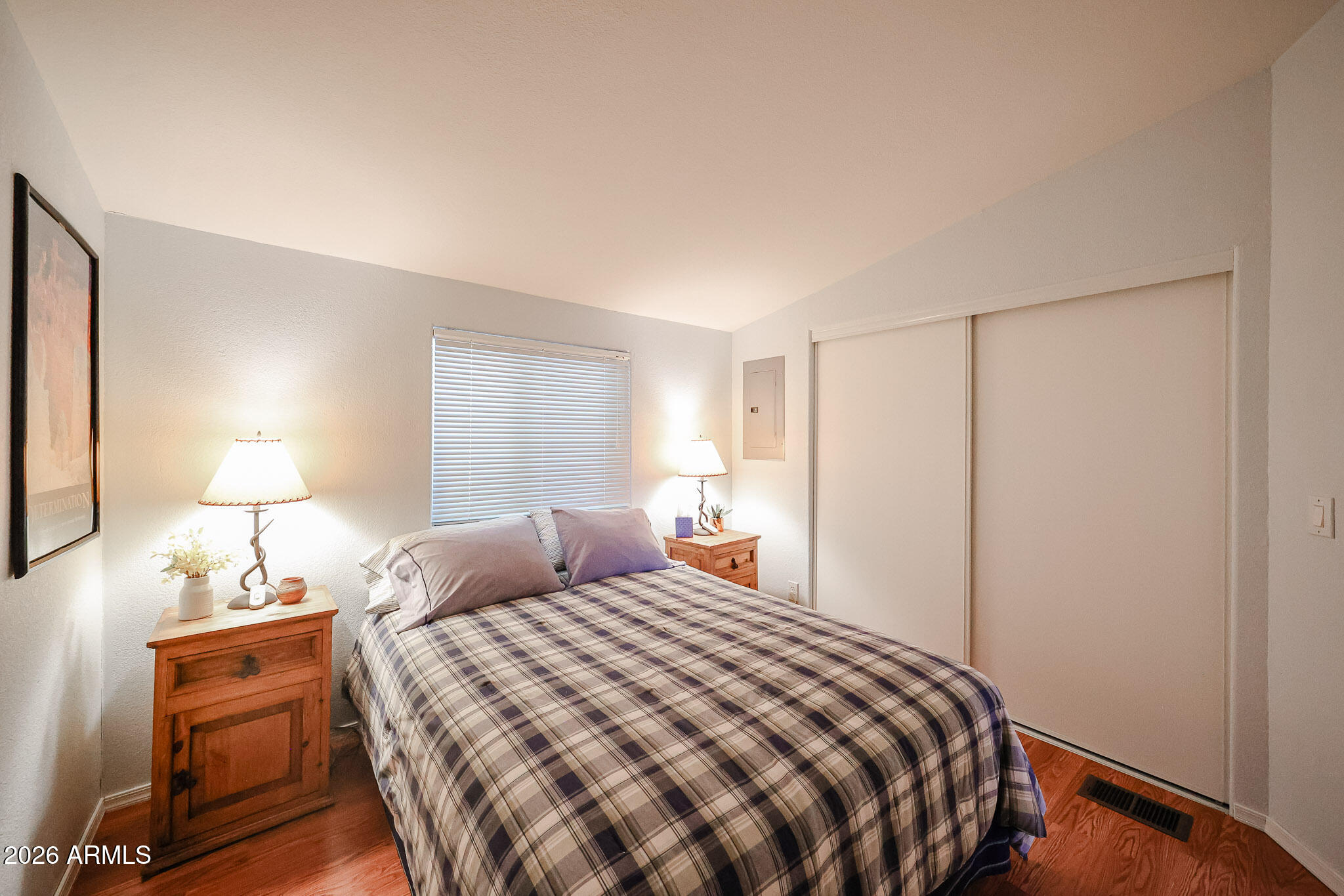 Furnished bedroom with a plaid-covered bed, wooden nightstands, window blinds, and sliding closet doors.
