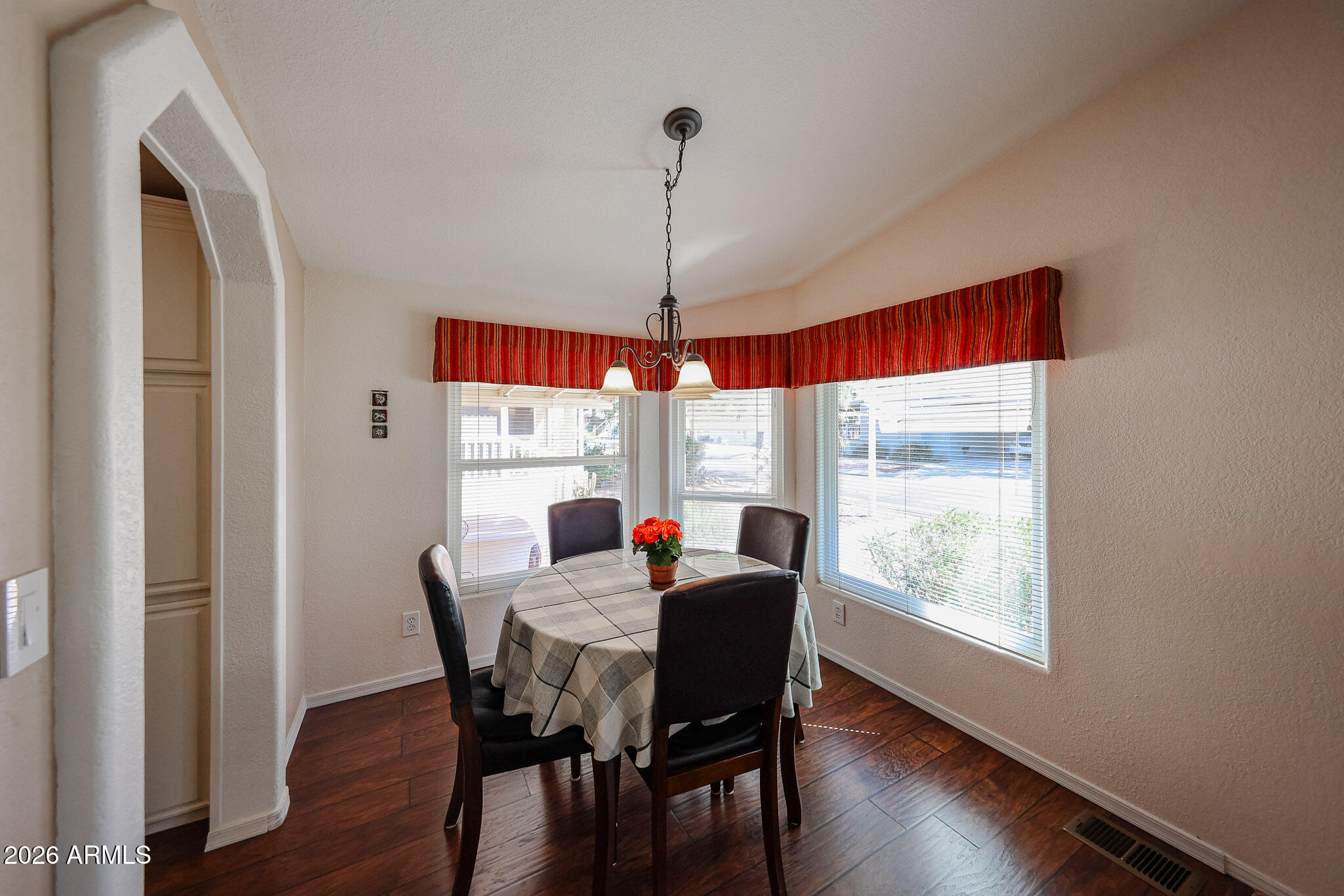 Dining nook with a checked tablecloth, dark chairs, three windows, and a red striped valance.
