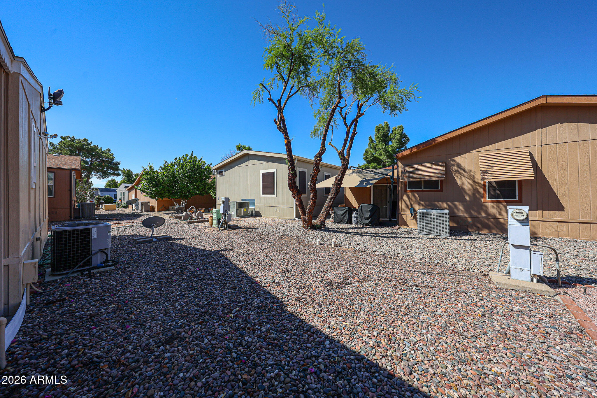 Gravel yard between single-story homes, with a mature tree and several air-conditioning units.