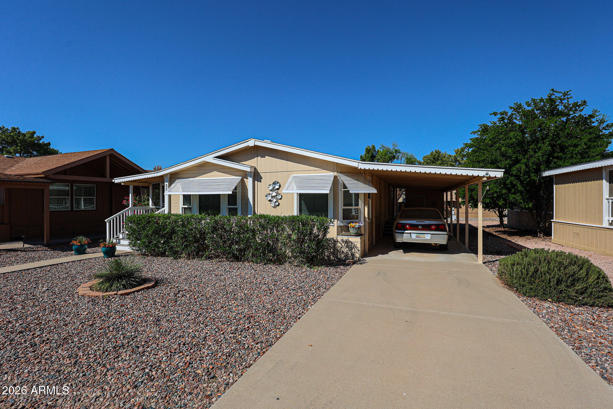 Beige single-story home with white awnings, gravel landscaping, and a car parked under its covered carport.