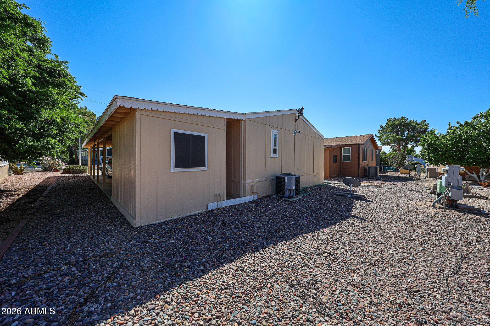 Tan manufactured home with a covered side porch and rock landscaping beside neighboring homes.