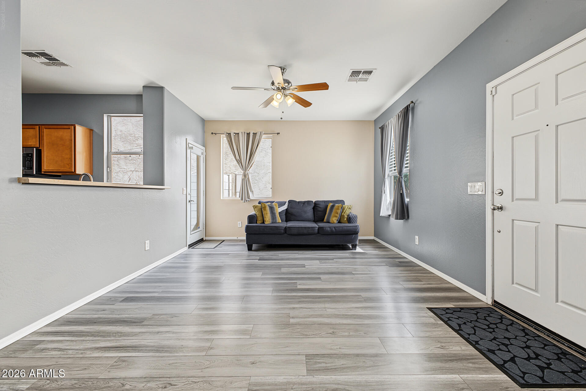 Living room with gray plank floors, a dark sofa, yellow accent pillows, and a kitchen pass-through.