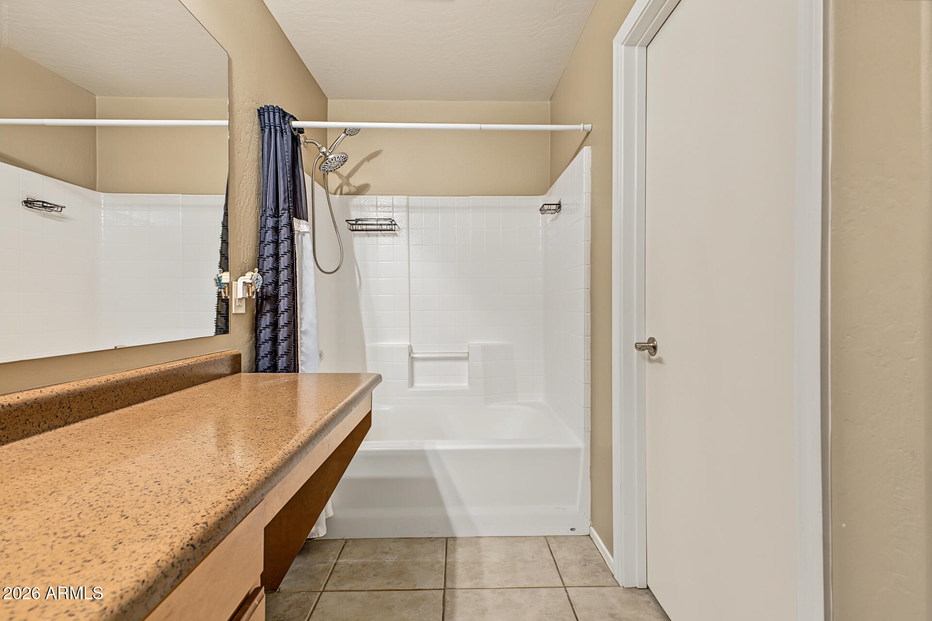 Bathroom with a white tub and shower, dark curtain, and tan-speckled counter beneath a large mirror.