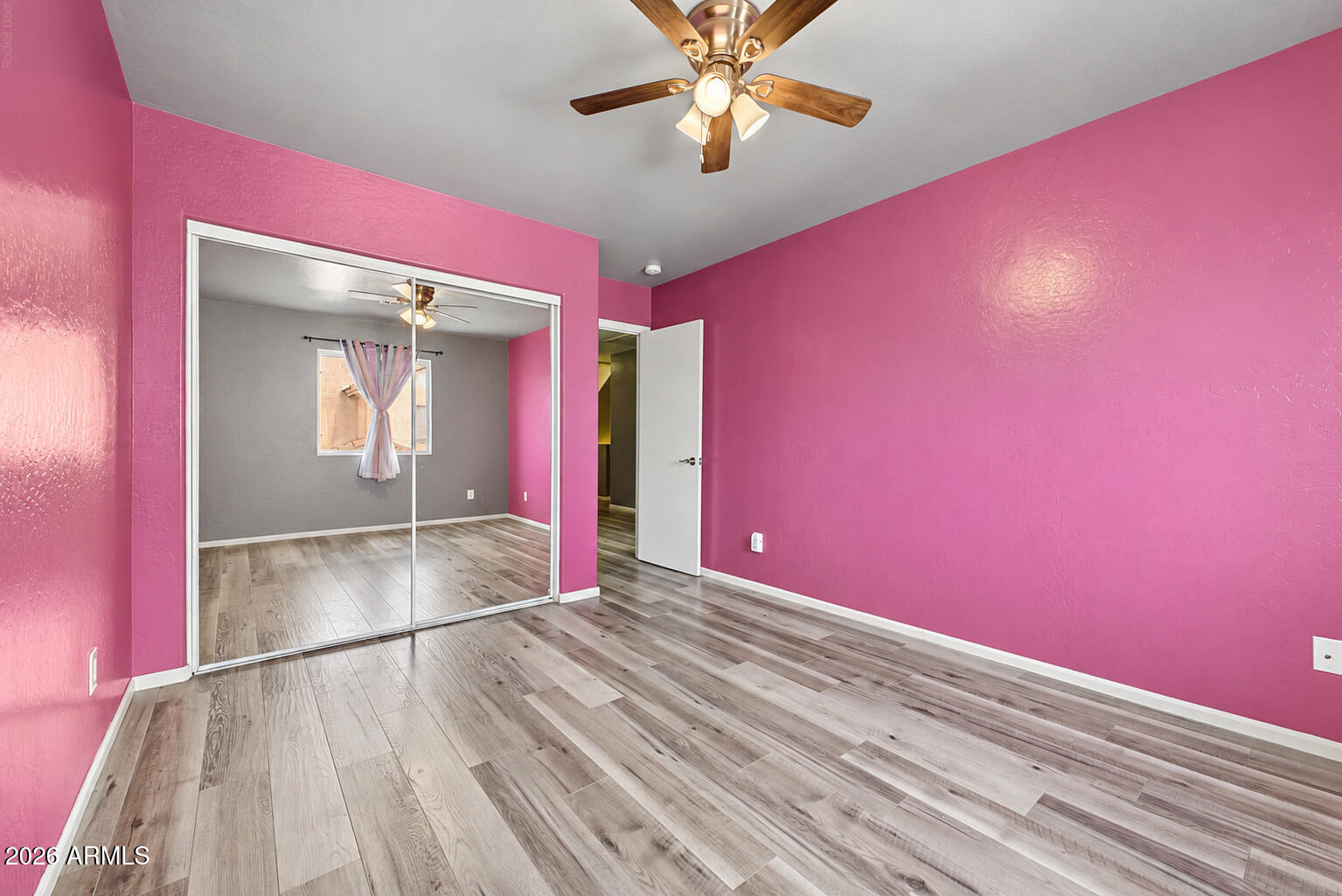Empty bedroom with bright pink walls, mirrored closet doors, gray wood-look flooring, and a ceiling fan.