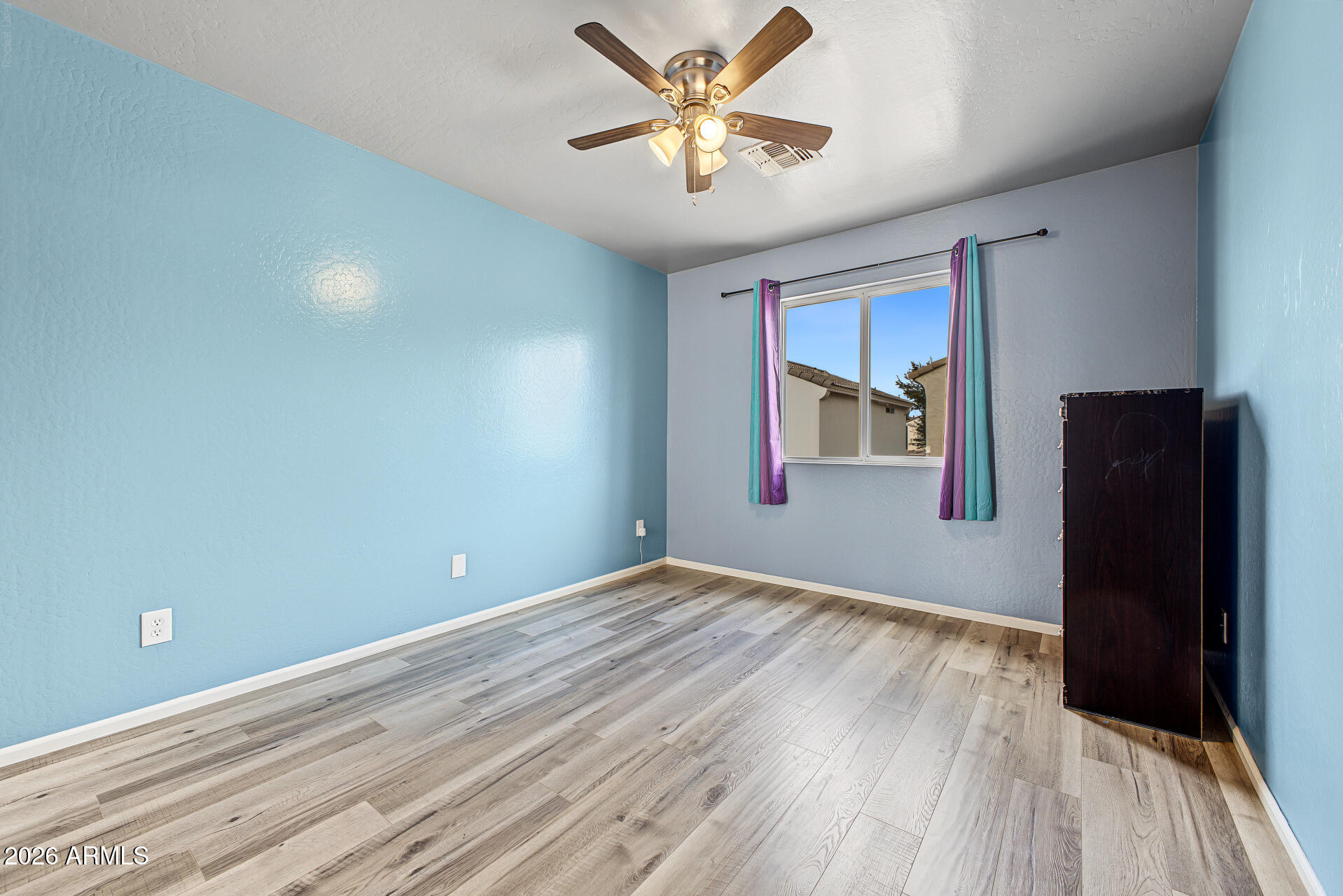 Empty bedroom with blue walls, plank flooring, a ceiling fan, and colorful curtains.