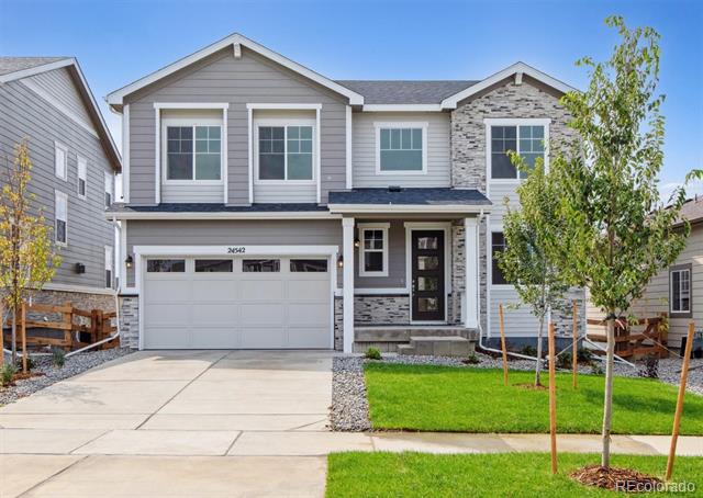 Two-story gray house with stone accents, a two-car garage, and a young tree beside the lawn.