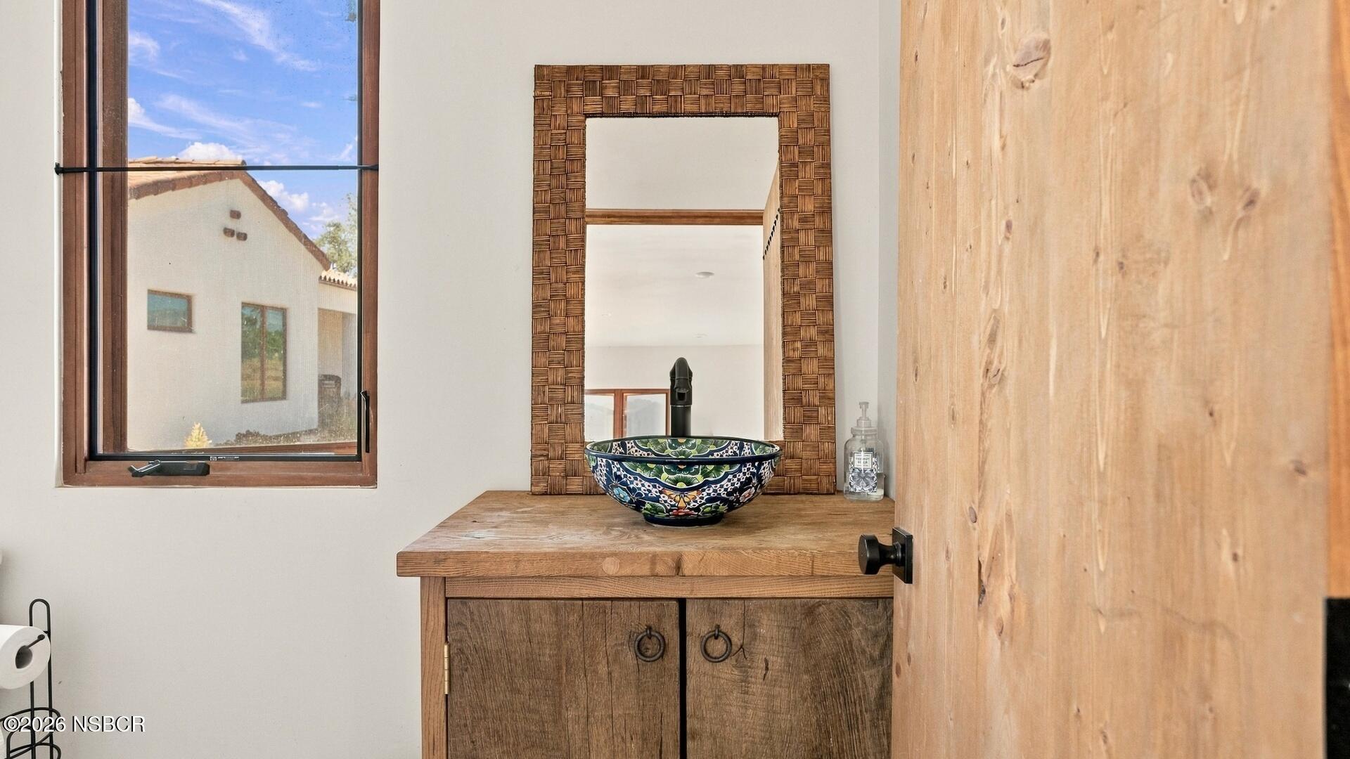 Bathroom vanity with a colorful patterned vessel sink, rustic wooden counter, and woven-frame mirror.