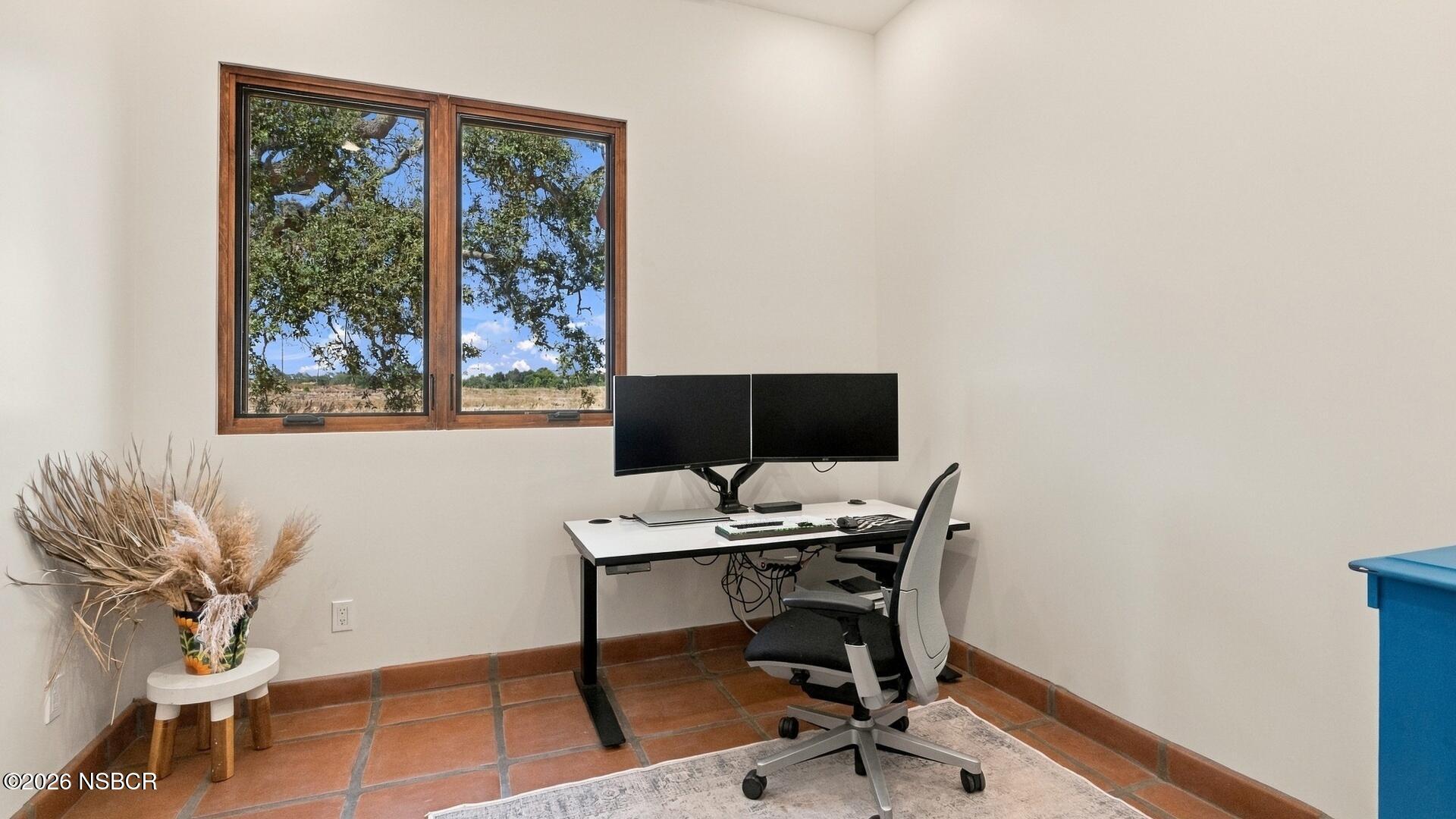Home office with dual monitors, terracotta tile floors, and a wood-framed window overlooking open countryside.