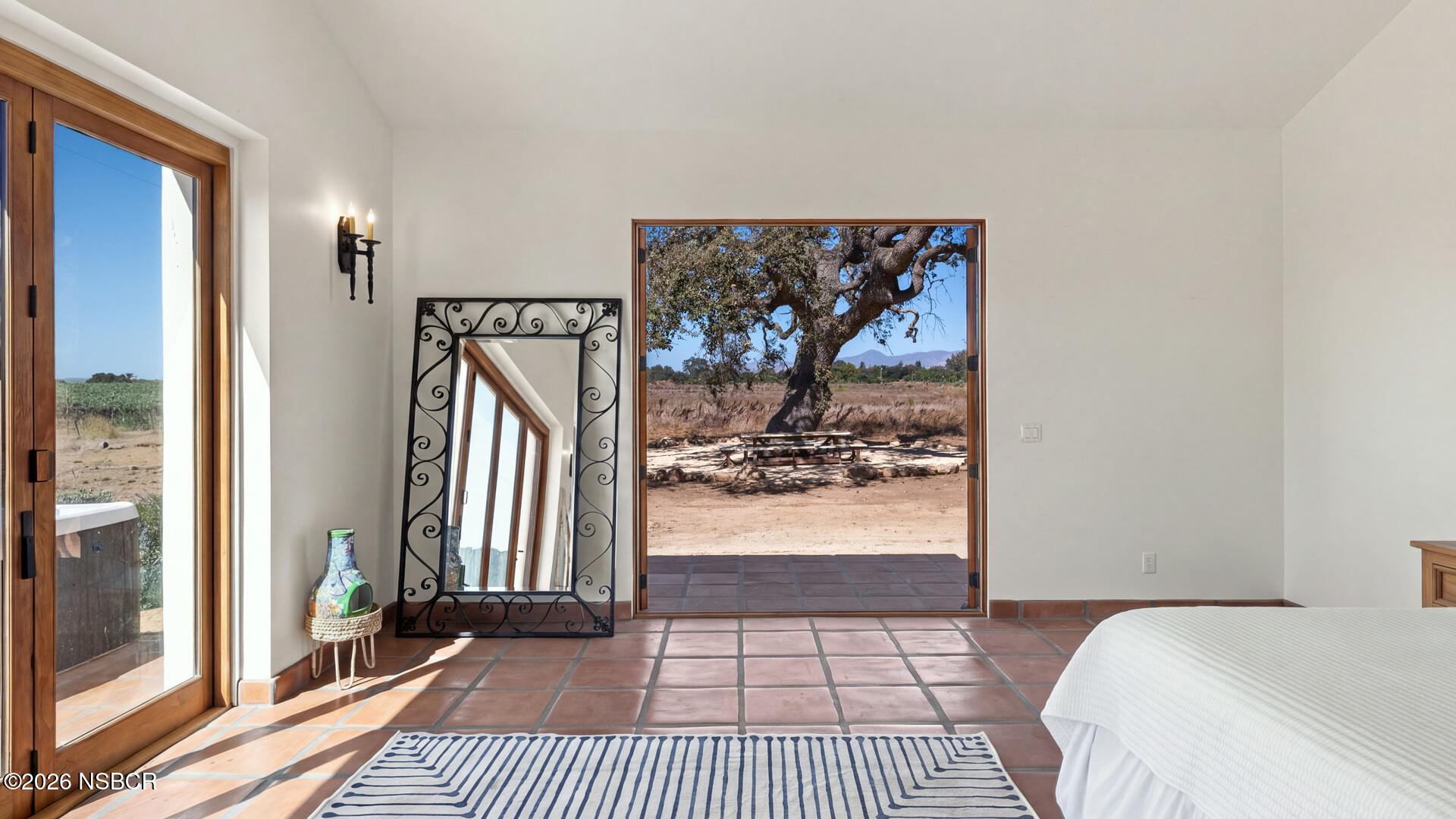 Bedroom with terracotta tile floors and open doors overlooking a vineyard and oak tree.