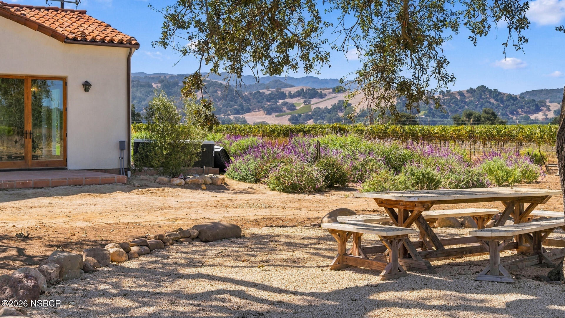 Vineyard-side patio with wooden picnic tables beside a stucco home, overlooking rolling hills.