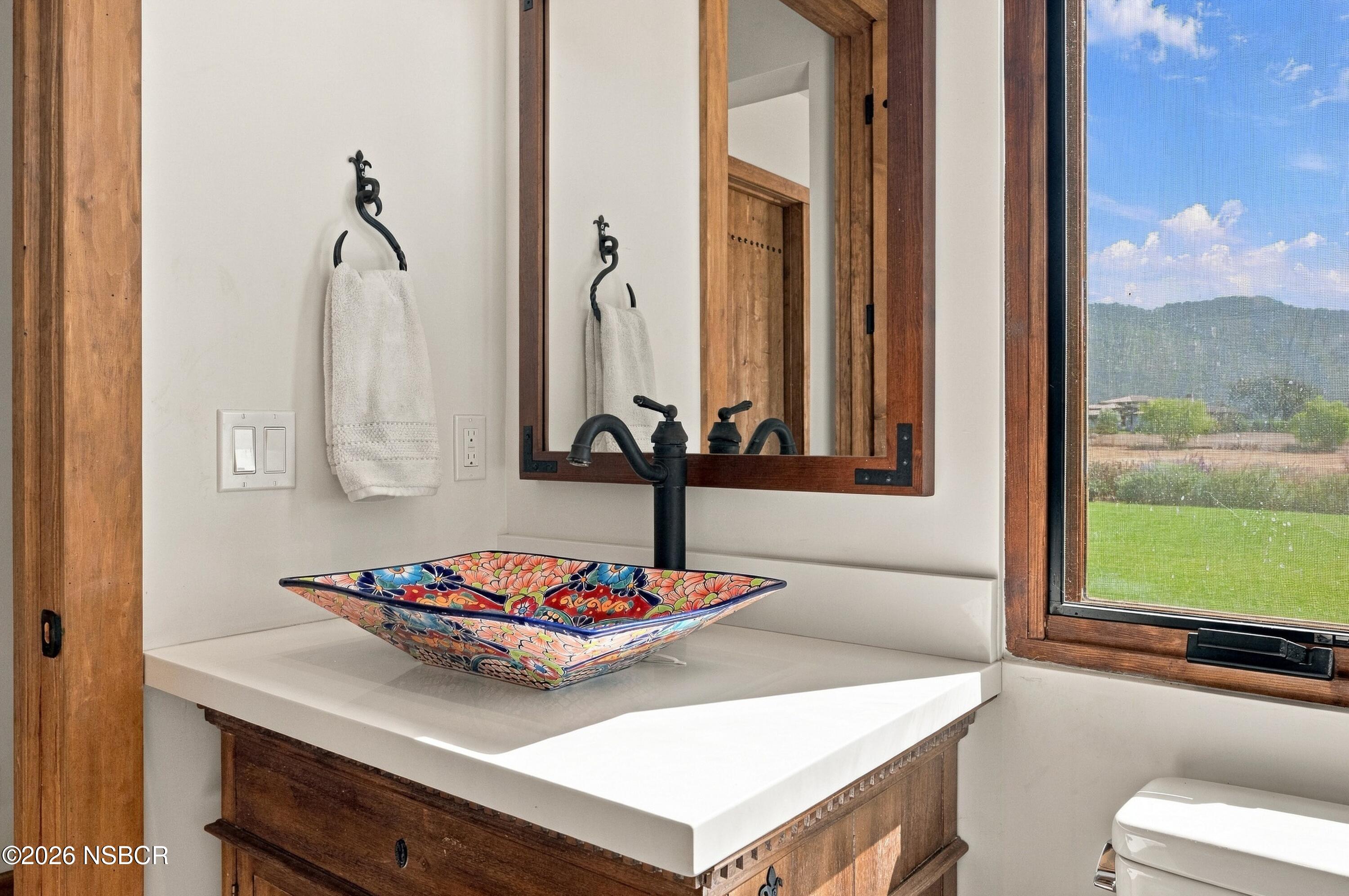Bathroom vanity with a colorful floral vessel sink, wood cabinetry, and a window overlooking a green lawn.