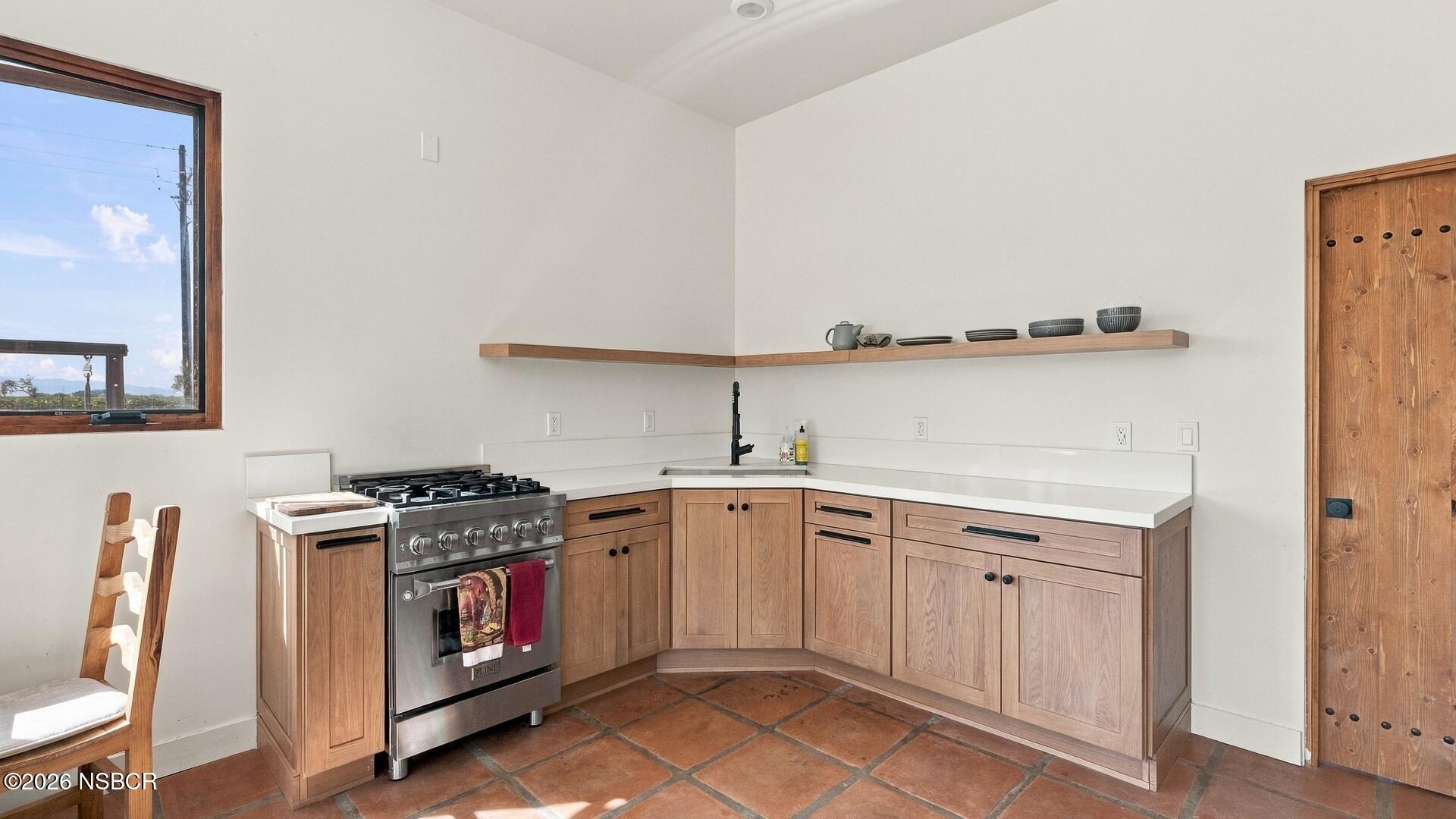 Kitchen with pale wood cabinetry, terracotta tile floors, and a stainless-steel gas range.