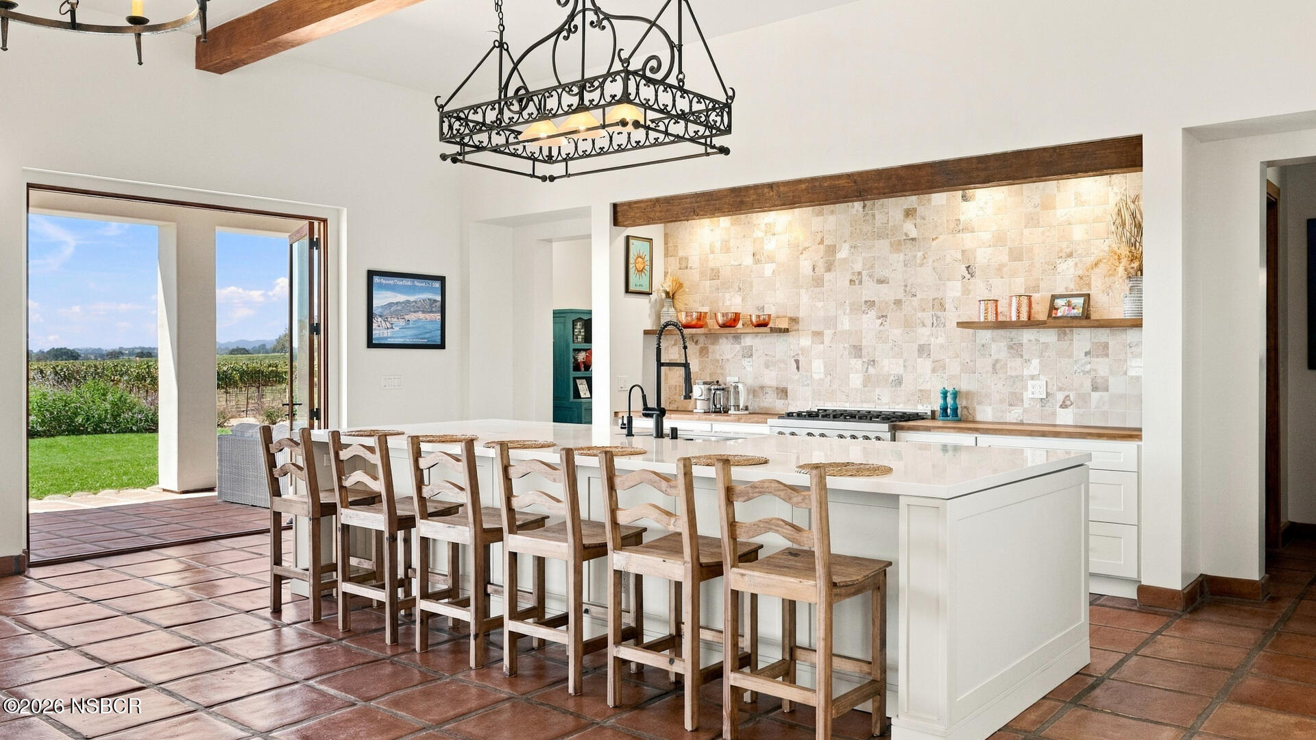 Kitchen with a large white island, wooden bar stools, and open doors overlooking a vineyard.