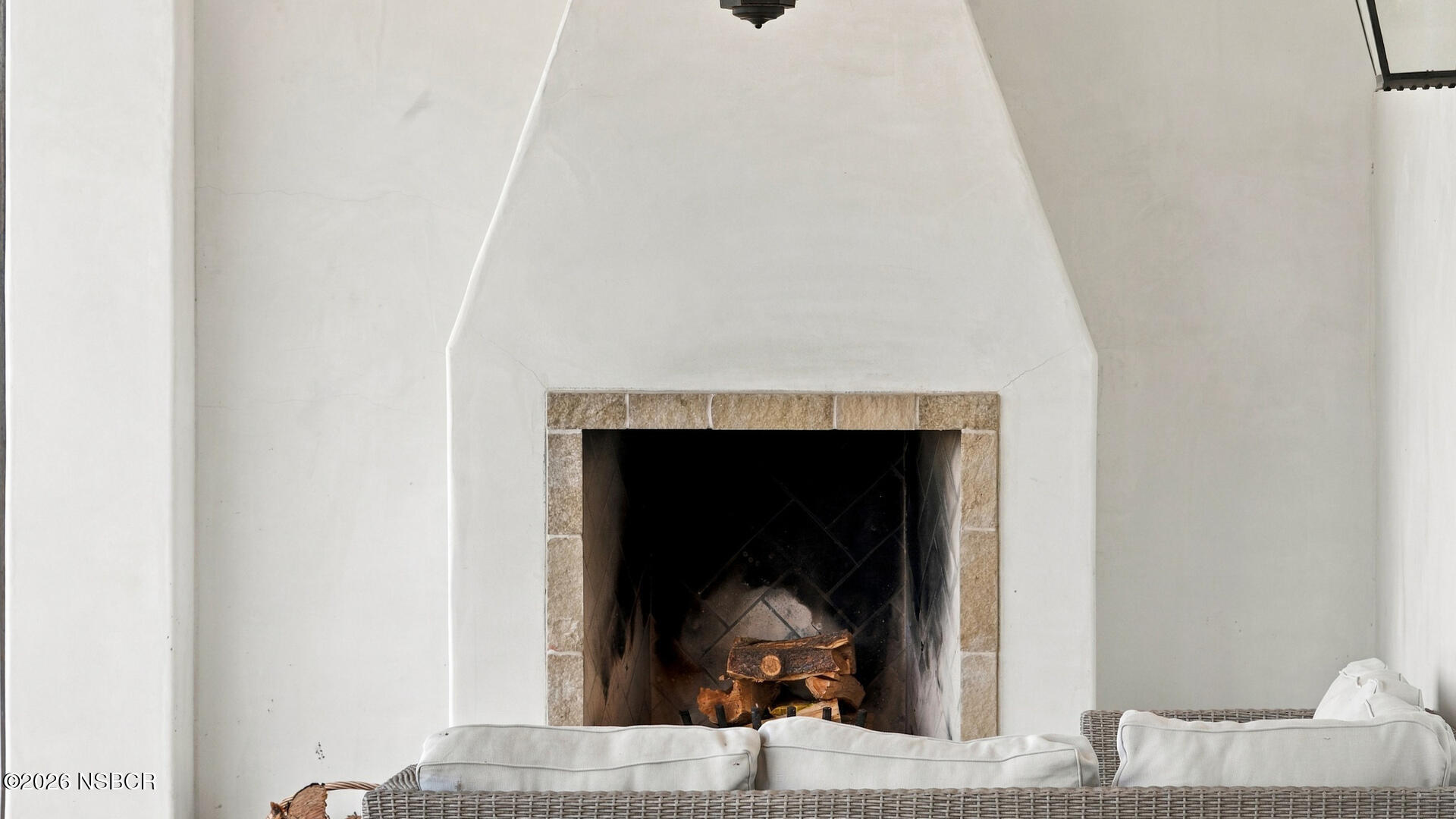 White plaster fireplace with tan stone trim and wicker seating in front.