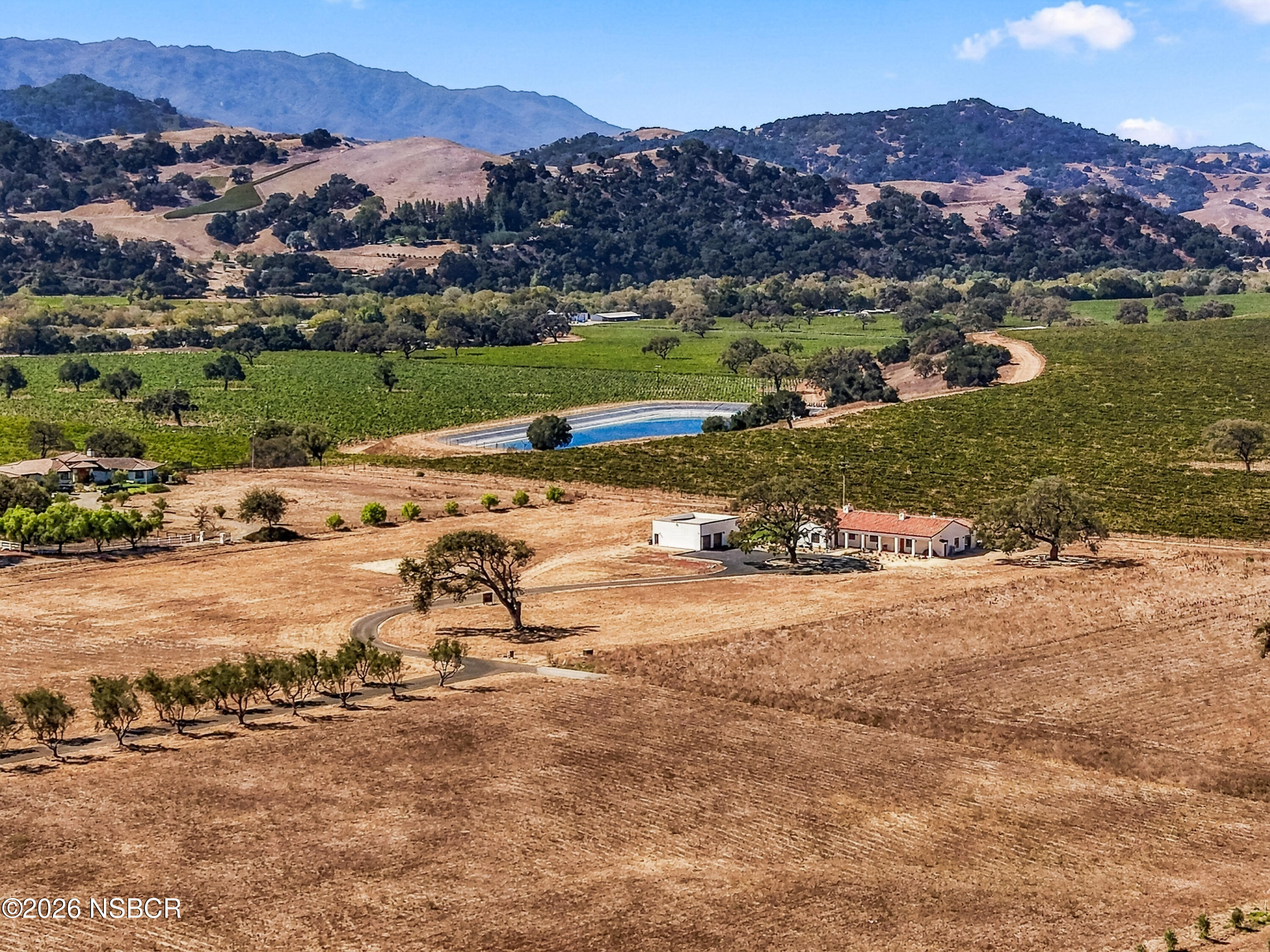 Aerial view of a ranch home and outbuilding amid dry fields, green vineyards, a blue pond, and rolling hills.