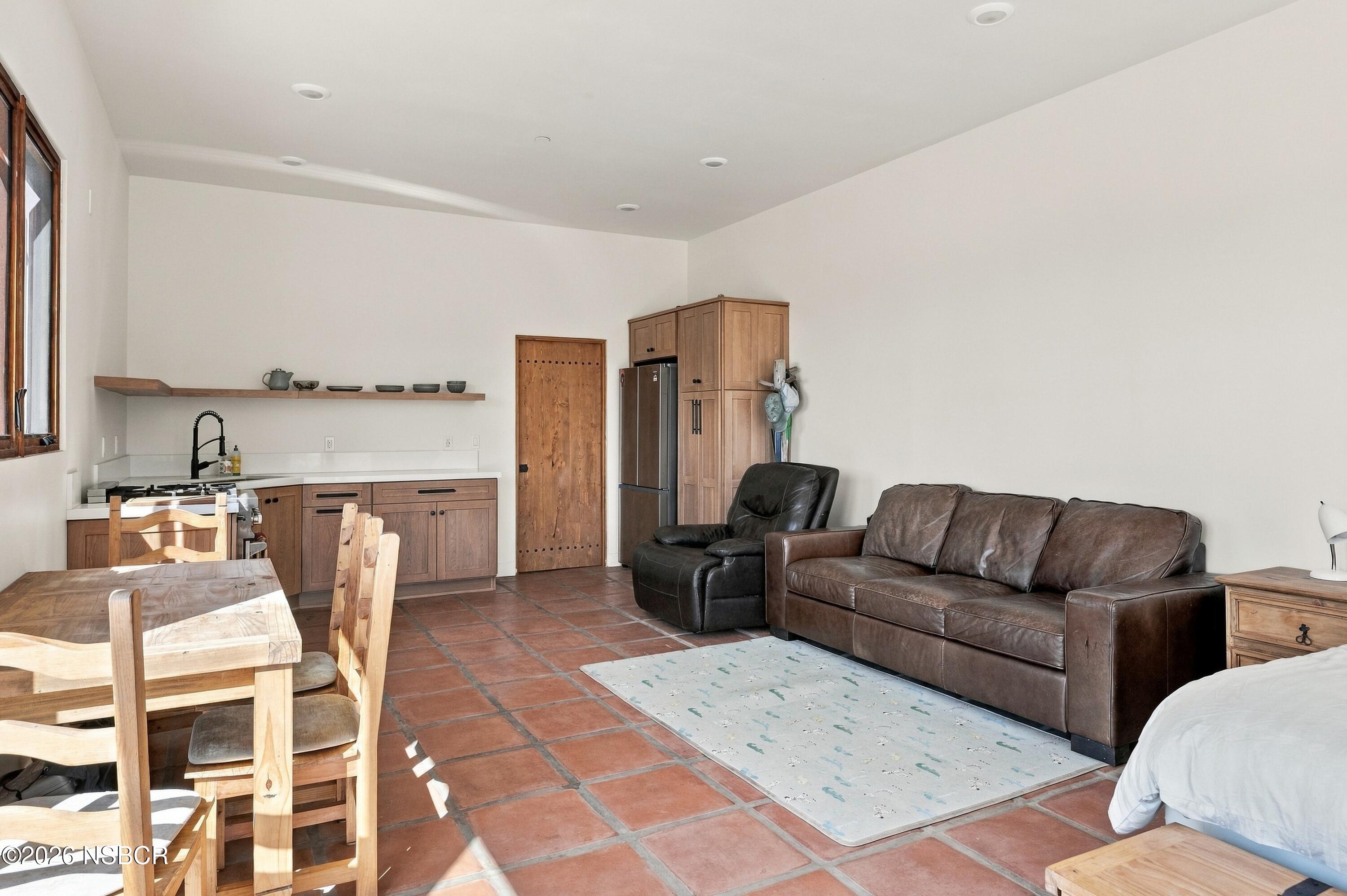 Open-plan living room with a compact kitchen, leather sofa, dining set, and terracotta tile flooring.