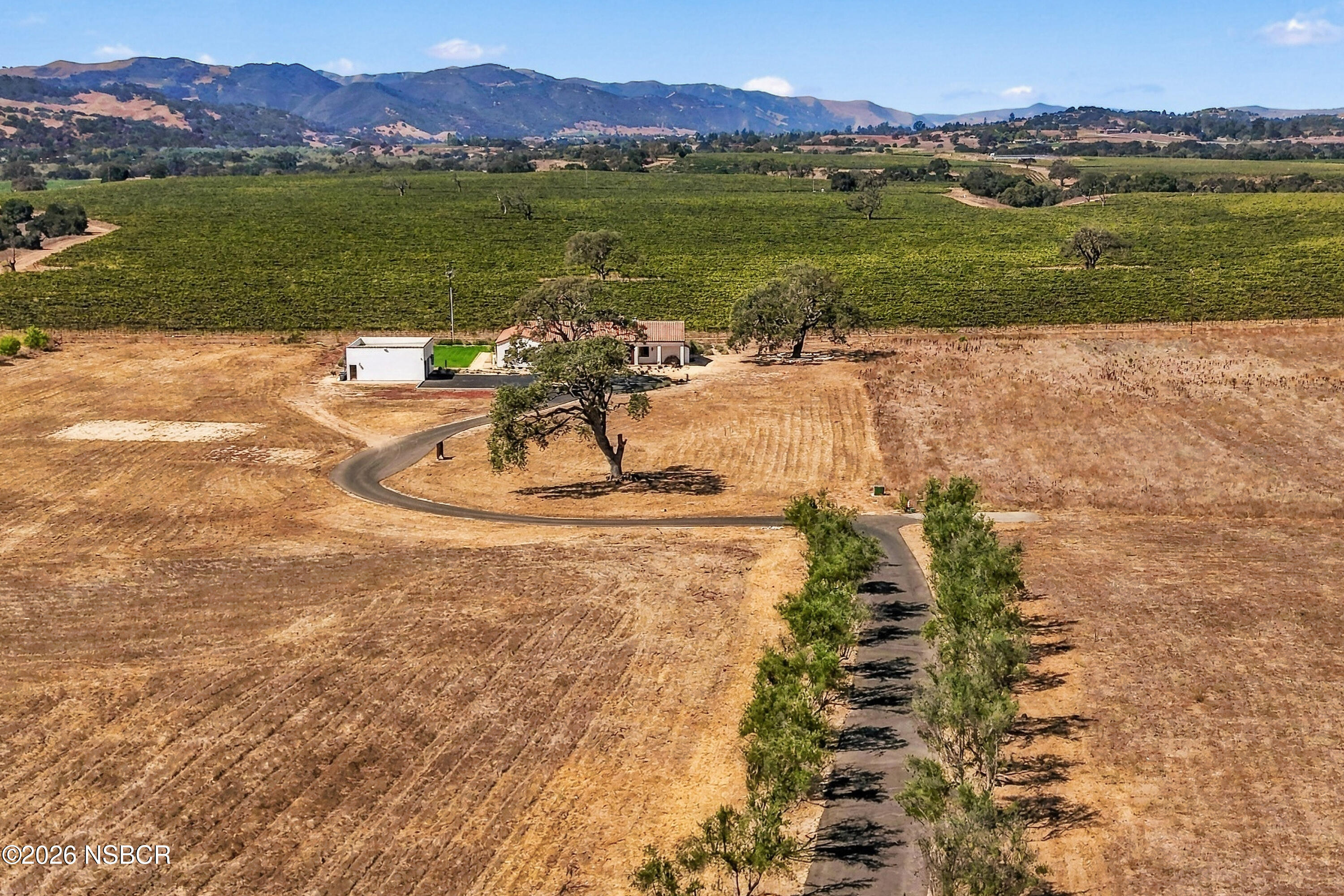 Aerial view of a rural home and outbuilding beside a curving driveway, with planted fields and mountains beyond.