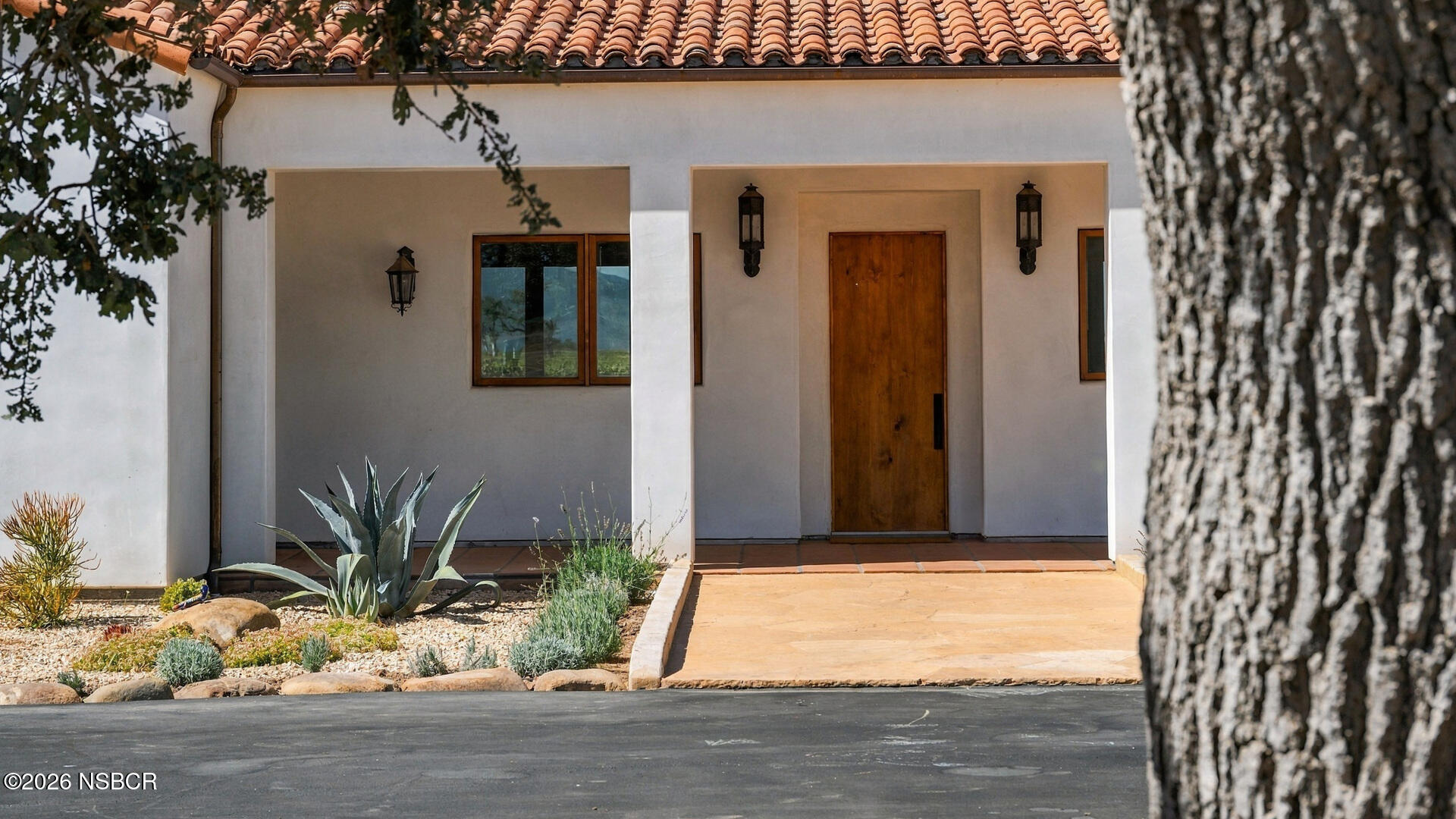 White stucco home entrance with terracotta tile roof, wooden door, and drought-tolerant landscaping.
