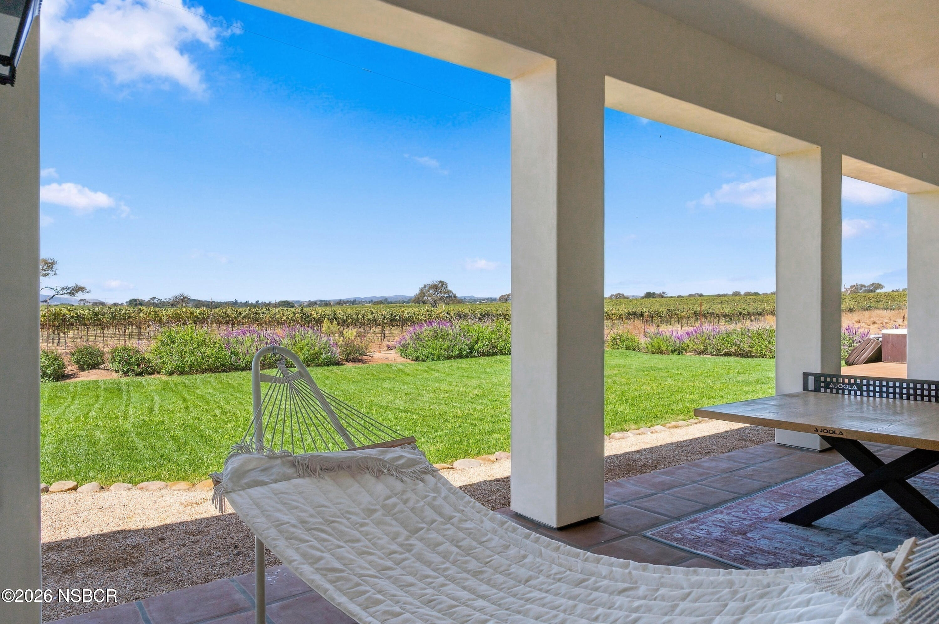 Covered patio with a hammock and ping-pong table overlooking a lawn and vineyard.
