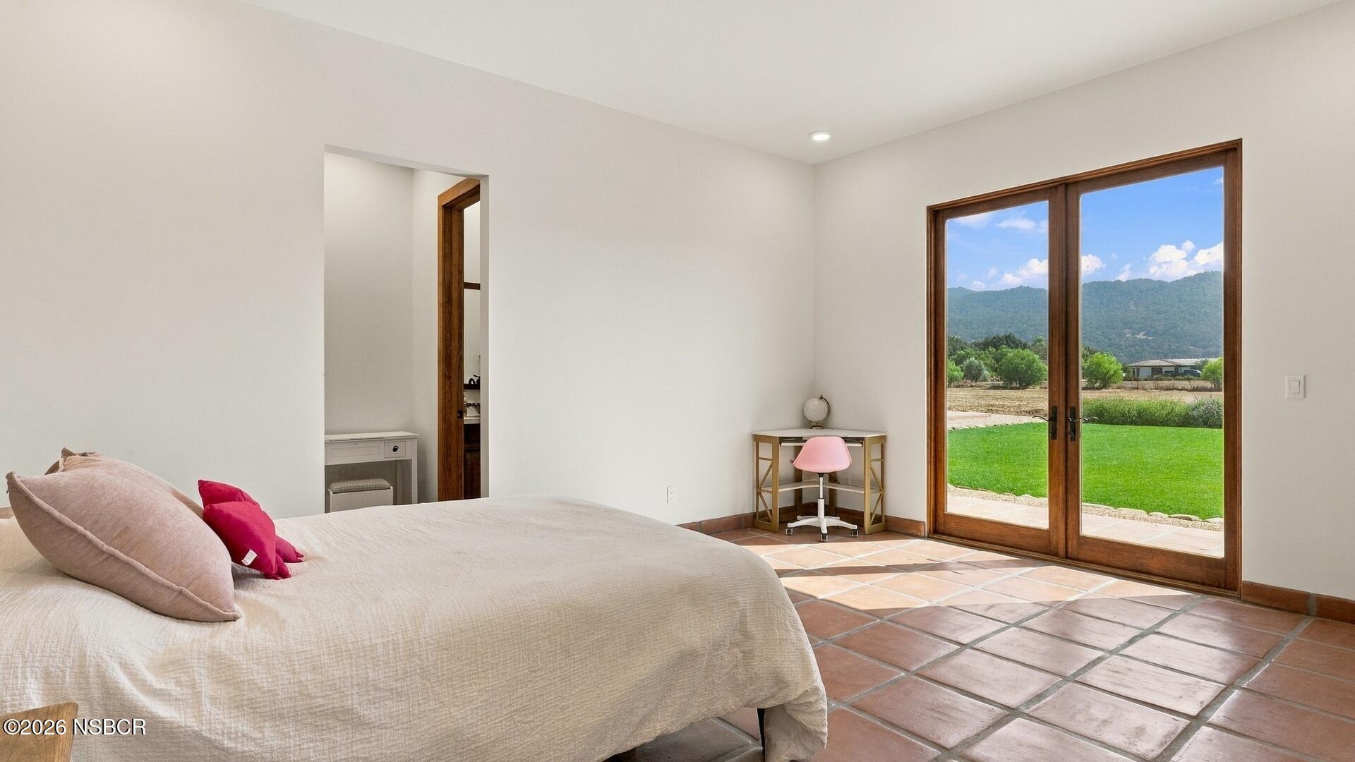 Bedroom with terracotta tile floors and wood-framed glass doors overlooking a lawn and green hills.