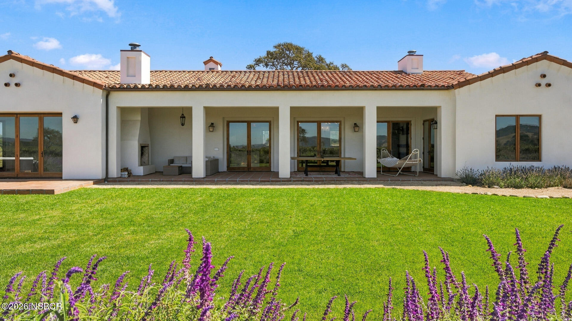 Stucco home with a covered patio, terracotta roof, green lawn, and purple flowers.