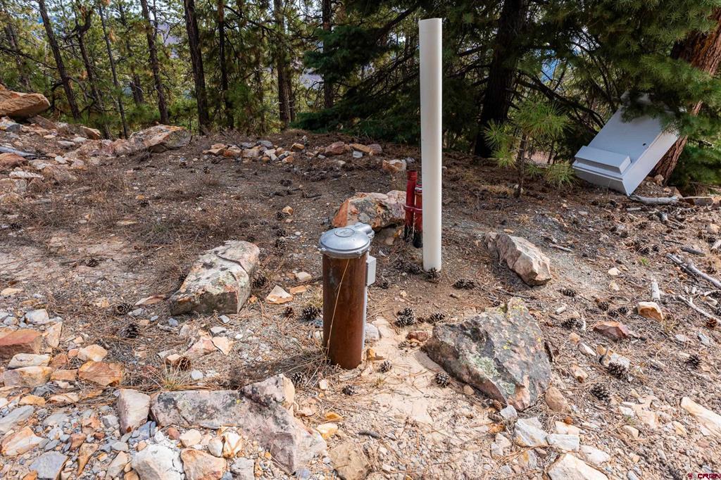 Rocky wooded lot with a capped pipe, tall white pipe, and metal utility cover.