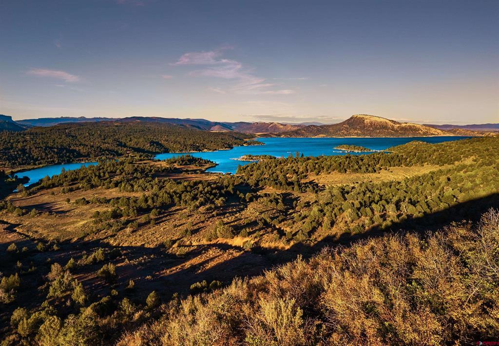 Blue lake amid evergreen forest and golden autumn hills, with mountains in the distance.