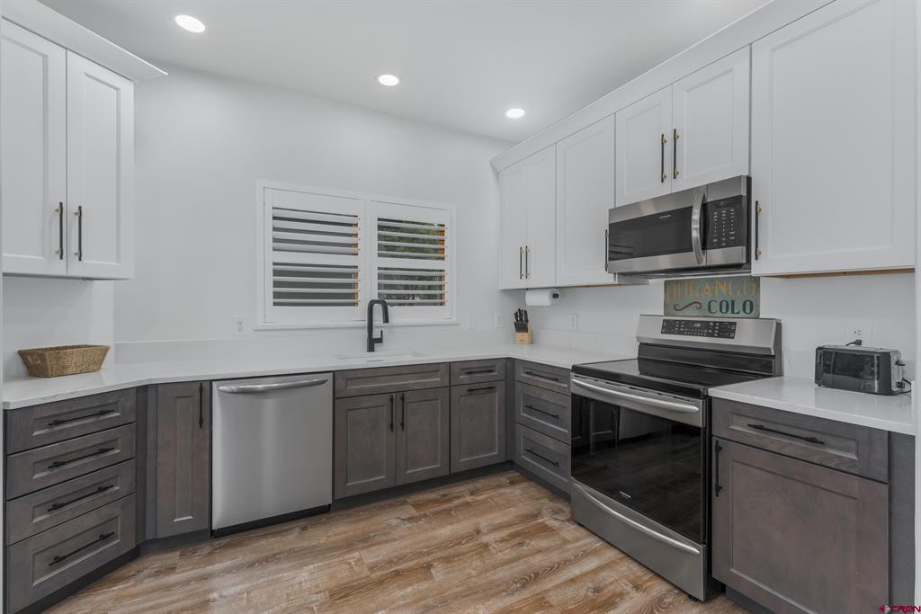 Modern kitchen with gray lower cabinets, stainless steel appliances, and a sink beneath a shuttered window.