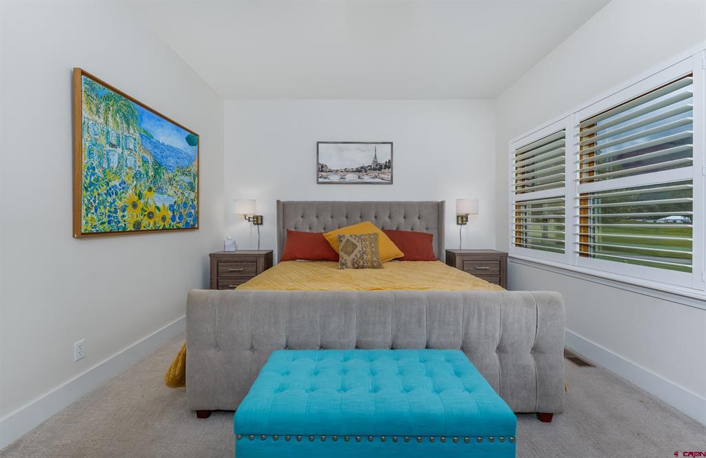 Furnished bedroom with yellow bedding, a blue bench, colorful artwork, and wide windows with blinds.