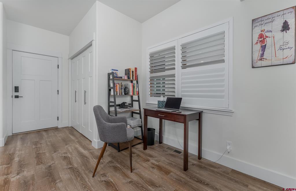 Home office with a wood desk, gray chair, ladder bookshelf, and white plantation shutters.