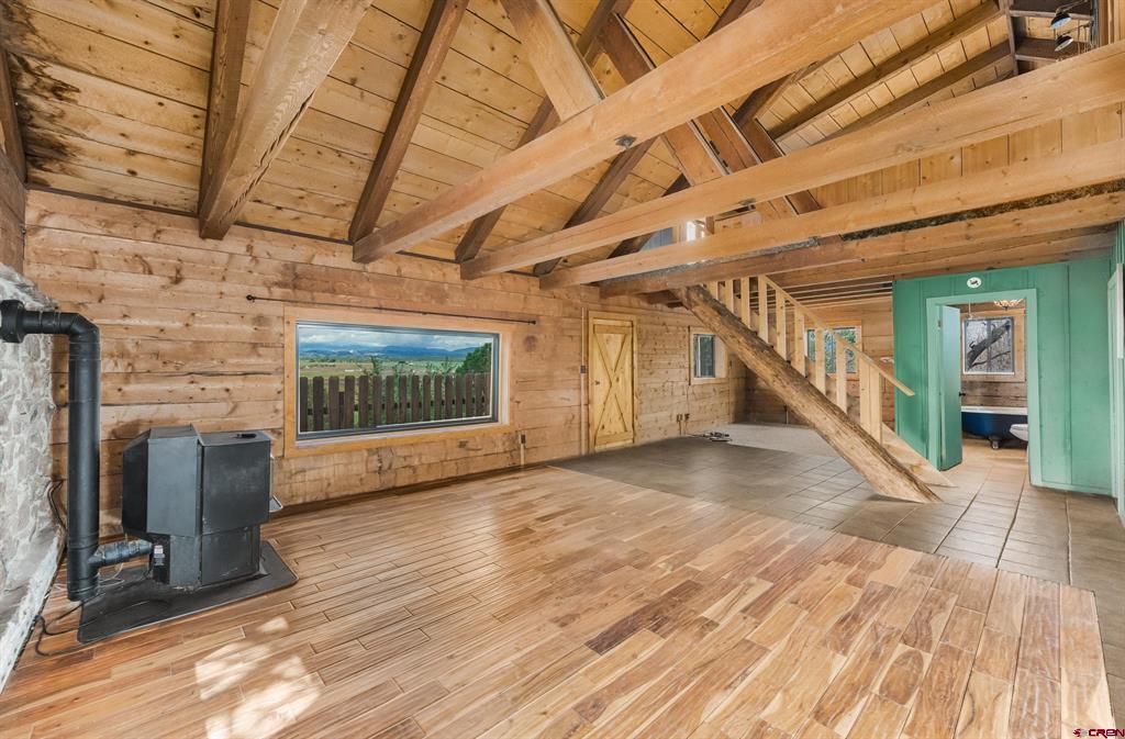 Rustic cabin interior with exposed timber beams, a black wood stove, and a window overlooking the countryside.