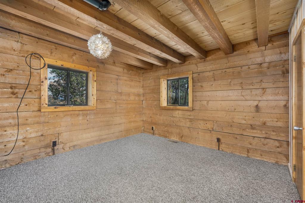 Empty wood-paneled room with exposed ceiling beams, gray carpet, and two windows.