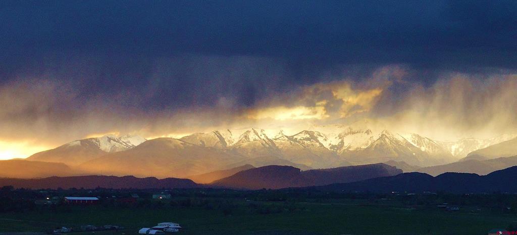 Snow-covered mountains glow above a rural valley beneath dramatic dark clouds.